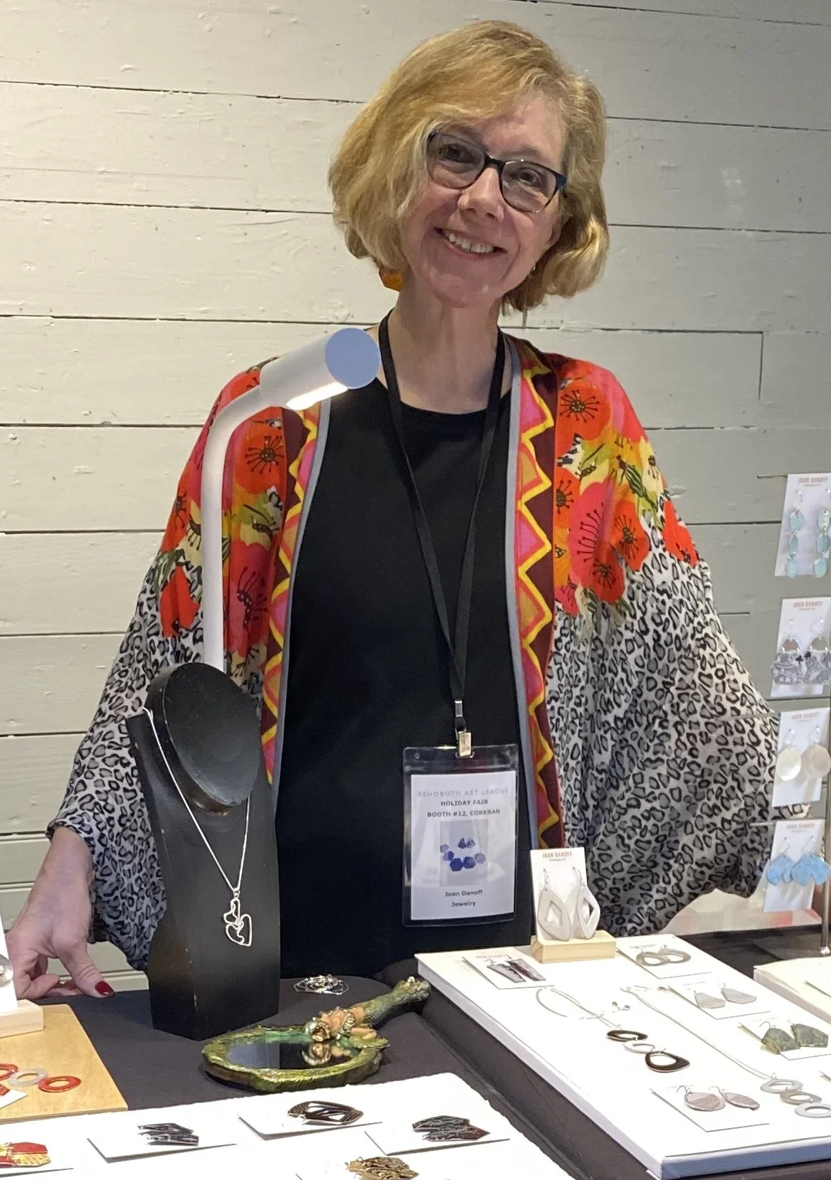 A woman with glasses and blonde hair smiling behind a jewelry display table at a craft fair, wearing a colorful floral and animal print shawl.