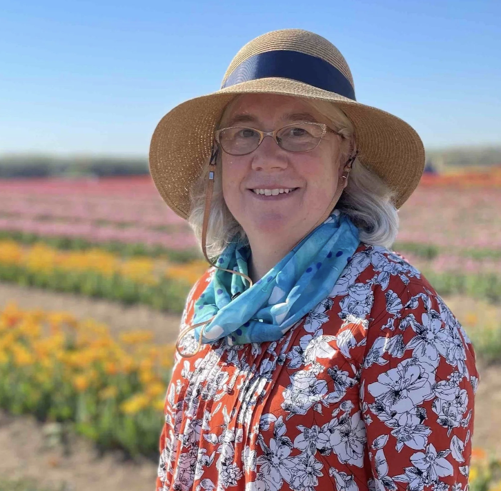 A woman smiling outdoors in a flower field, wearing a wide-brimmed straw hat with a black band, glasses, a red floral dress, and a blue scarf around her neck.