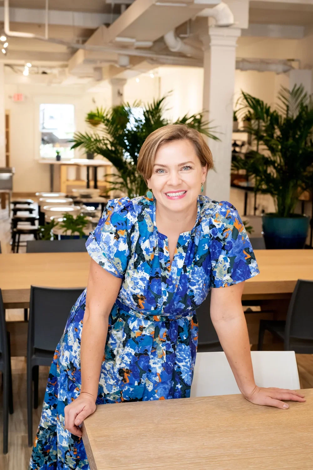 A woman in a blue patterned dress smiling and leaning on a table in a bright, modern restaurant or cafe with indoor plants and wooden furniture.