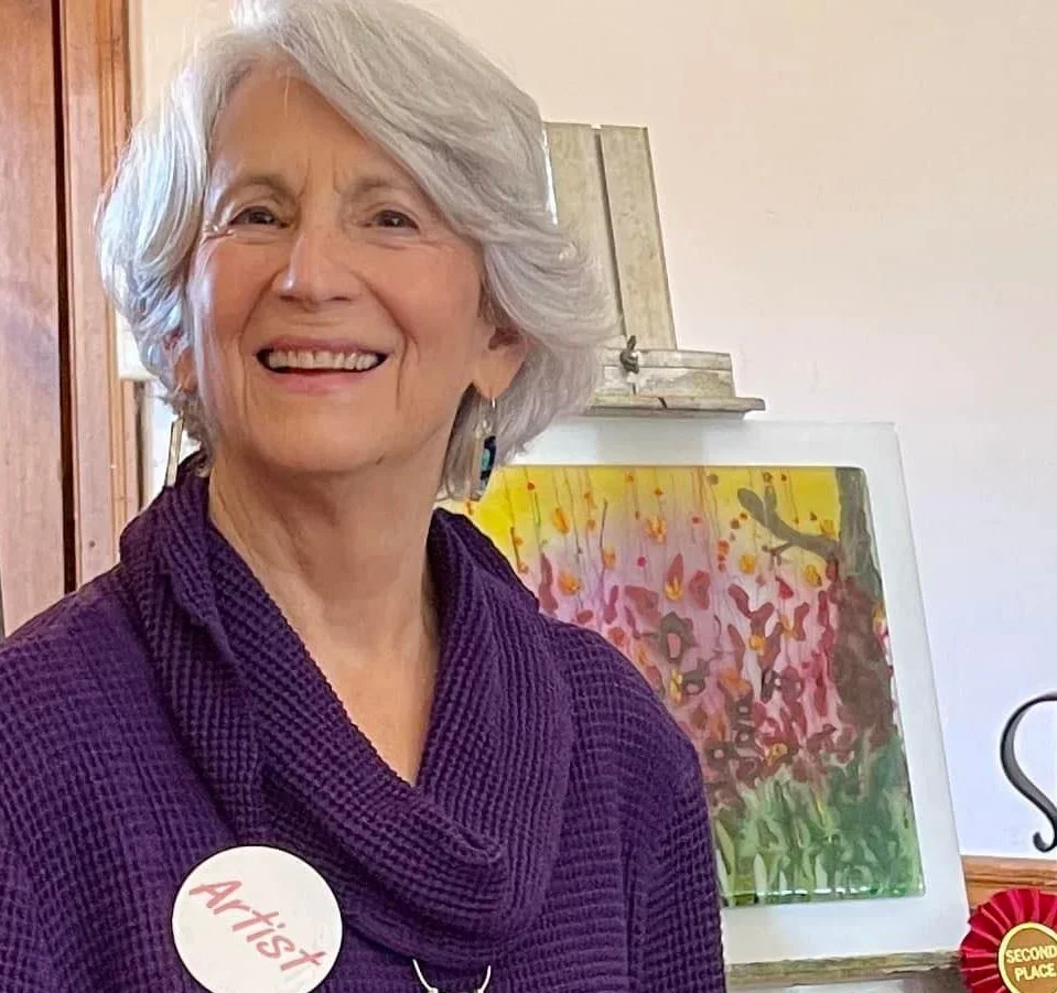 Smiling older woman with short white hair wearing a purple shawl or sweater, standing in front of colorful floral artwork, with a badge that says 'Artist' and a second-place ribbon.