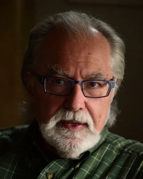Close-up photo of an older man with gray hair, glasses, and a white beard, wearing a green plaid shirt.