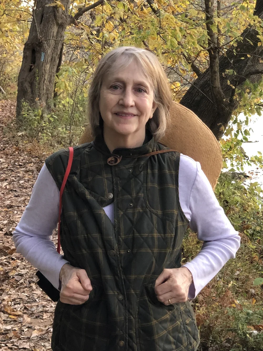 A woman with short gray hair smiling outdoors on a trail surrounded by autumn yellow and green leaves, wearing a plaid vest over a white long-sleeve shirt and carrying a woven hat on her back.