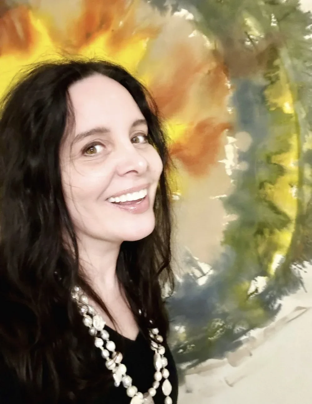 A woman with long dark curly hair smiling in front of a colorful abstract painting with yellow, orange, green, and blue hues.