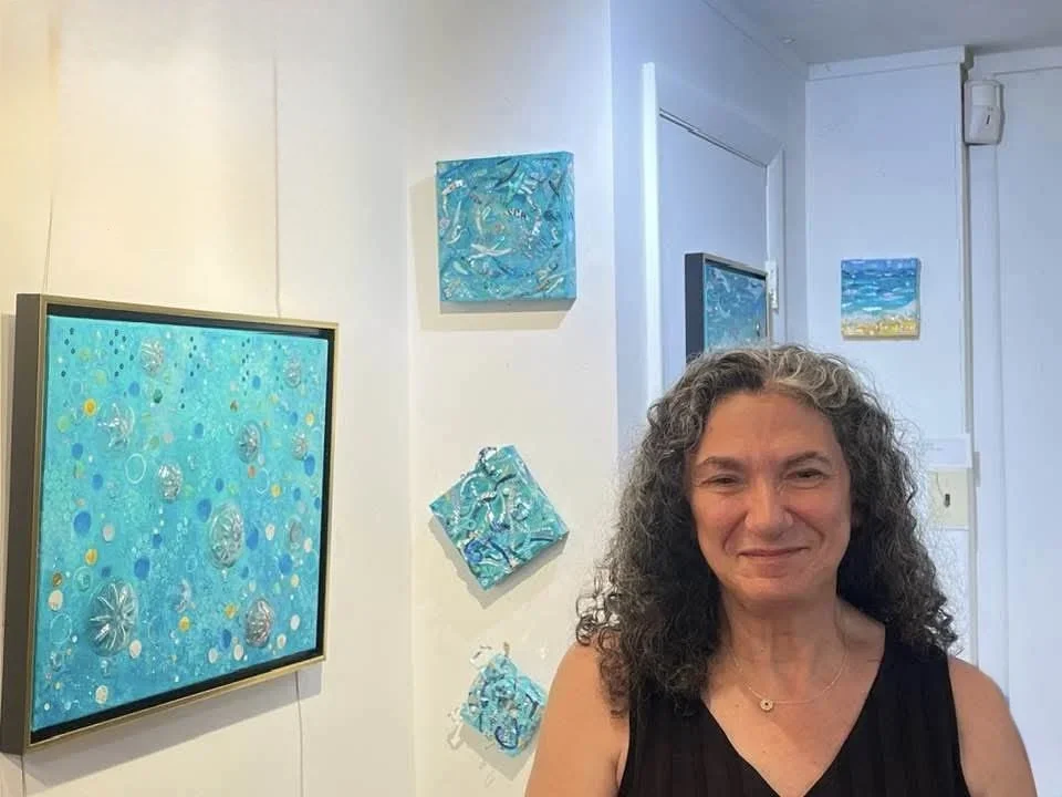 A woman with curly gray hair smiling at an art gallery with abstract blue paintings on the wall.
