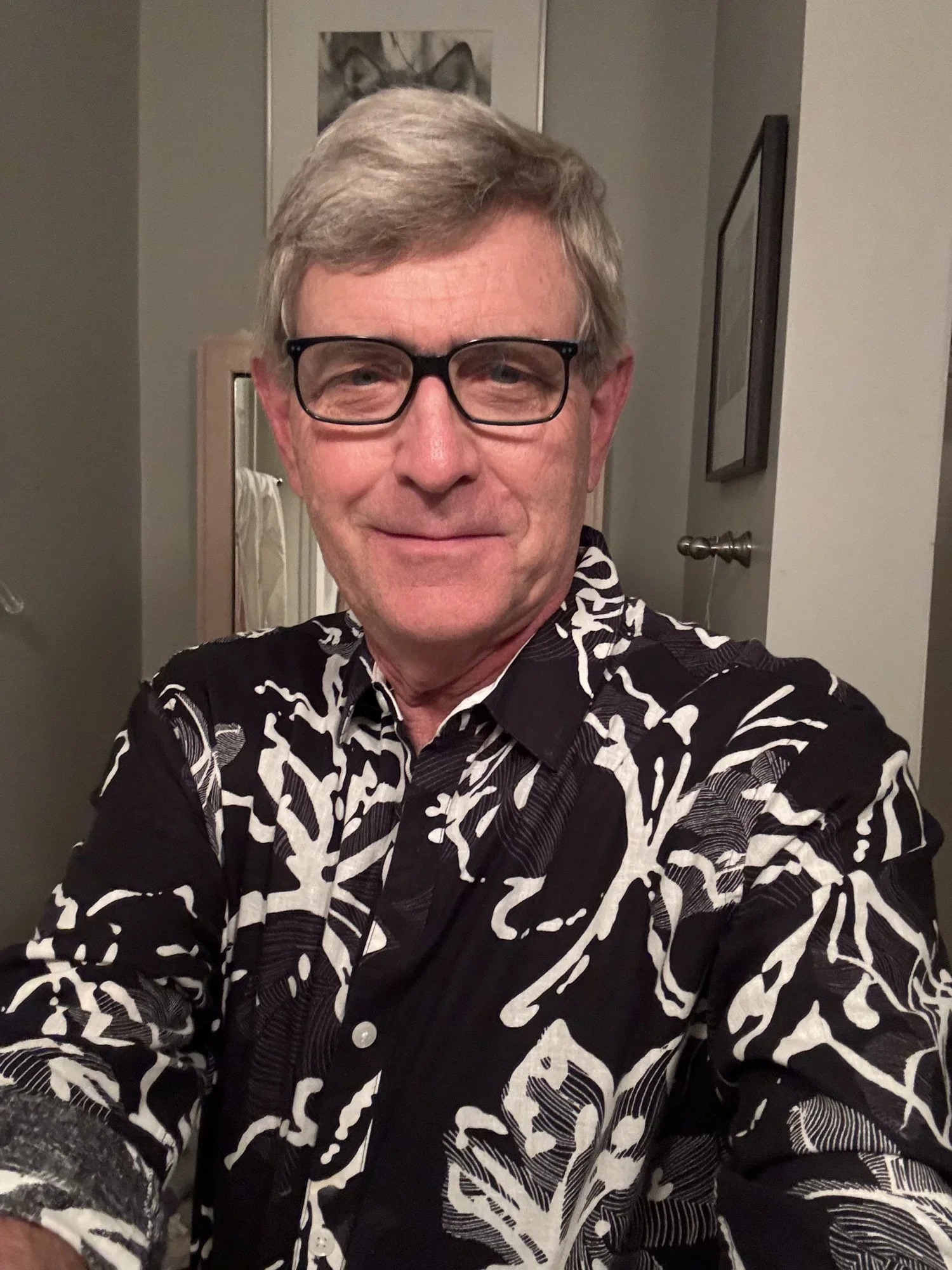 A man with gray hair and glasses taking a selfie in a room with a dresser mirror, a framed picture of a horse, and a wall-mounted picture of a butterfly in the background. He's wearing a black shirt with a white abstract pattern.