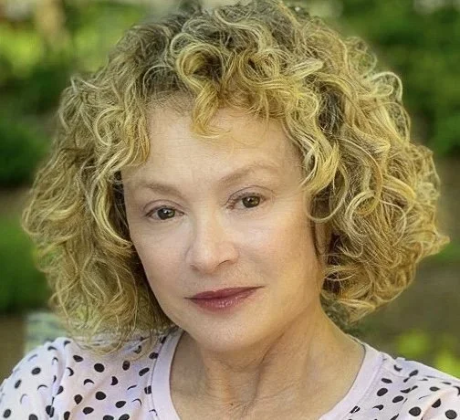 Close-up of a woman with curly blonde hair, wearing a polka-dotted top, outdoors with greenery in the background.