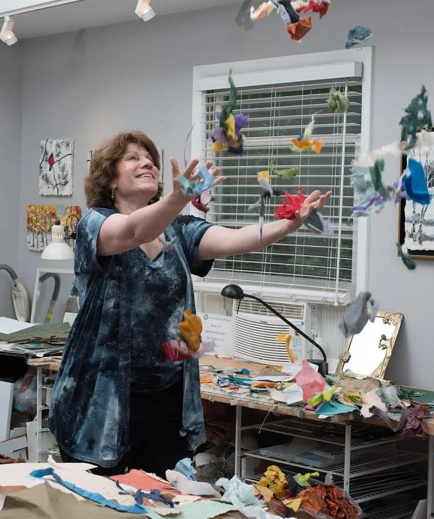 A woman standing in a craft room, throwing colorful pieces of fabric or paper into the air with a joyful expression.