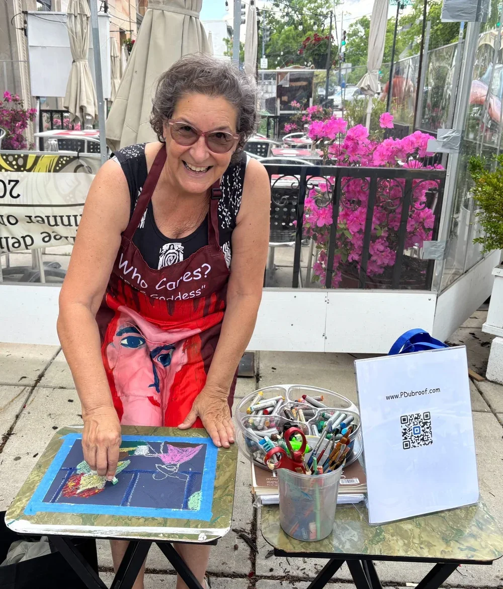 A smiling woman wearing sunglasses and a colorful apron is sitting at a table outdoors, working on a chalk art piece on a tray. There are art supplies like chalk and scissors on the table, and a QR code sign with a website URL next to her. Pink flowers and outdoor restaurant furniture are visible in the background.