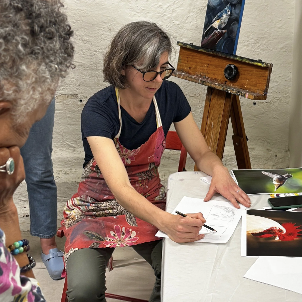 Woman drawing on paper at an art class with a painting of a bird on an easel behind her.
