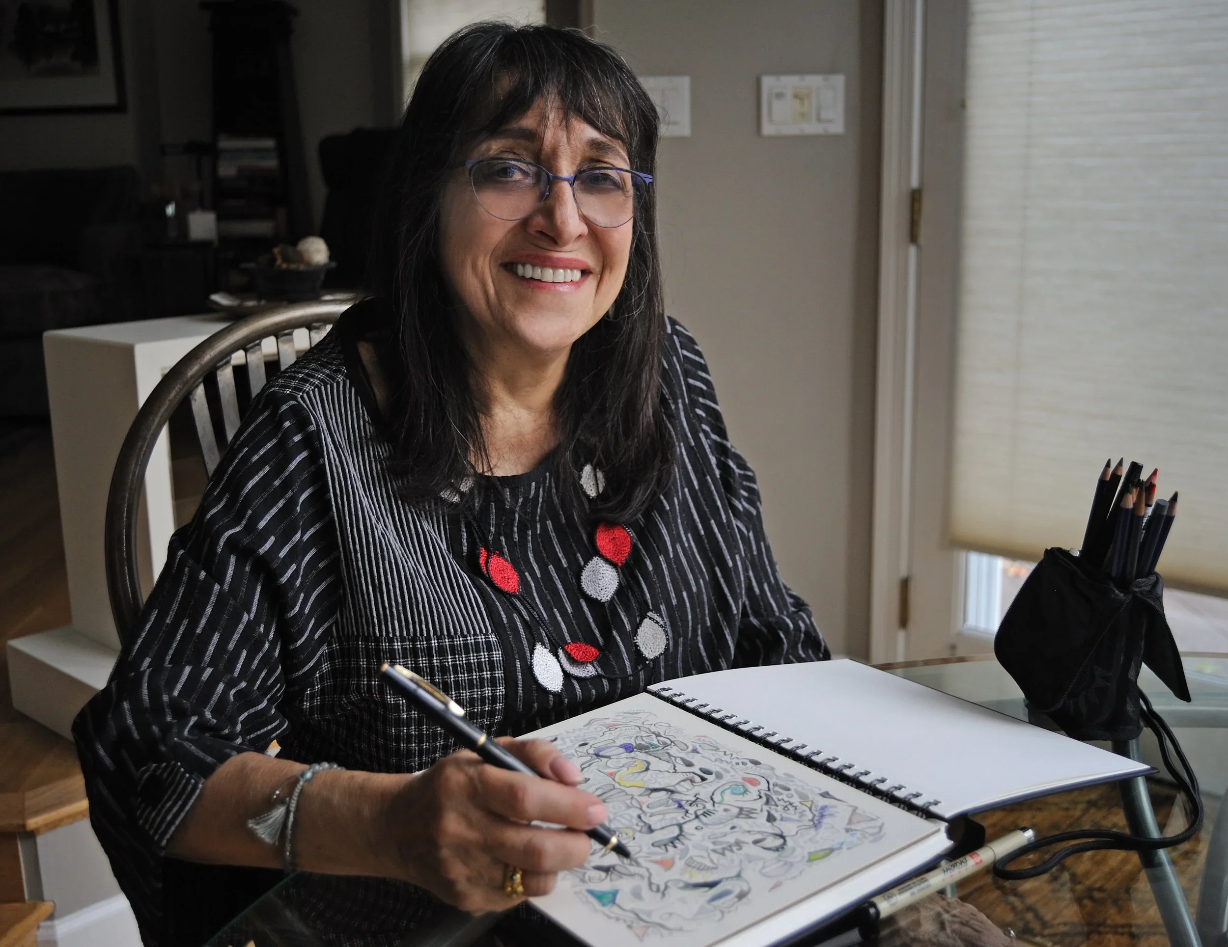 A middle-aged woman with dark hair, glasses, and a striped shirt is sitting at a table, drawing or coloring in a sketchbook with colorful markers.