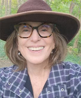 A woman outdoors wearing a wide-brimmed hat, glasses, and a plaid shirt, smiling at the camera.
