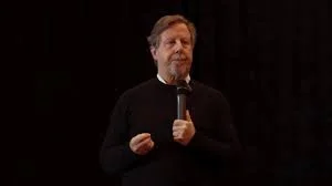 A man with a beard holding a microphone on stage, wearing a black sweater and a collared shirt, standing against a dark background.