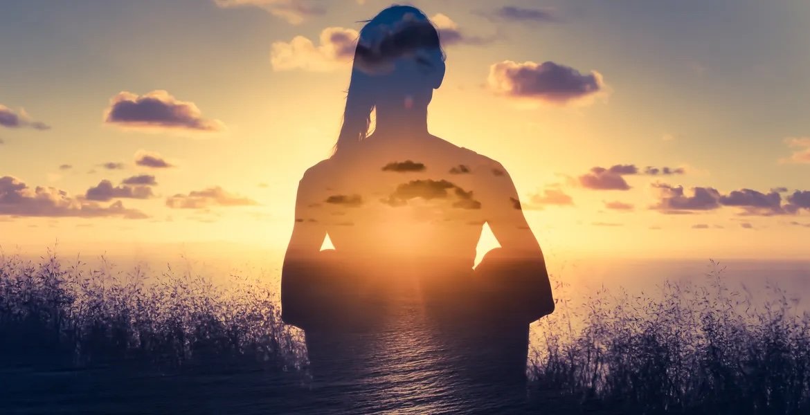 A double exposure photograph of a woman standing on a hillside during sunset, with her silhouette blending into a scenic view of clouds, the sky, and the ocean.