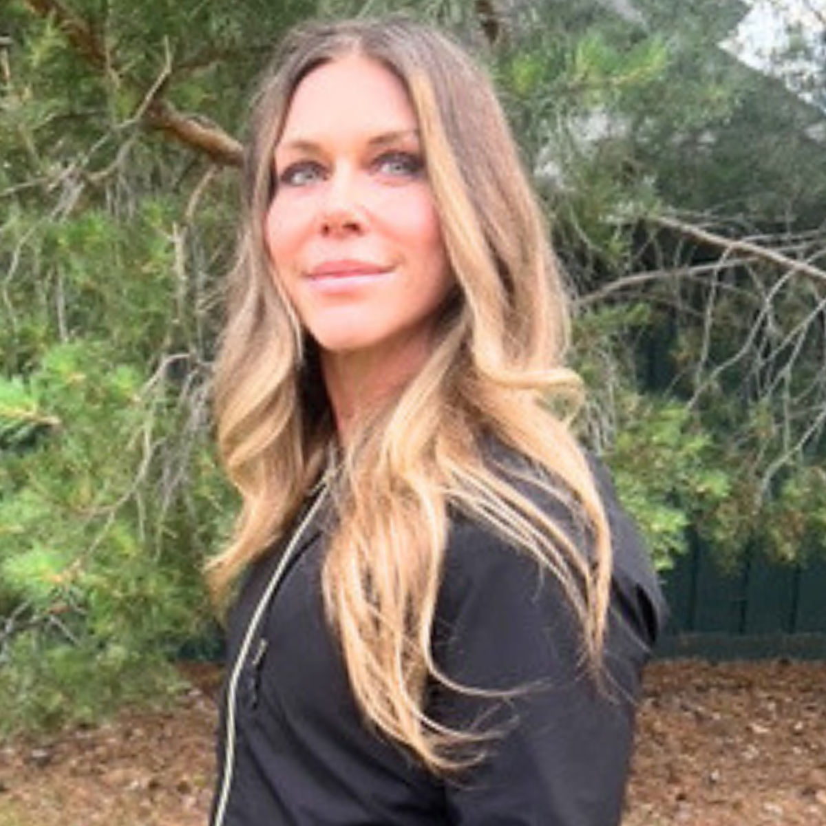 A woman with long wavy blonde hair and light skin standing outdoors near green trees