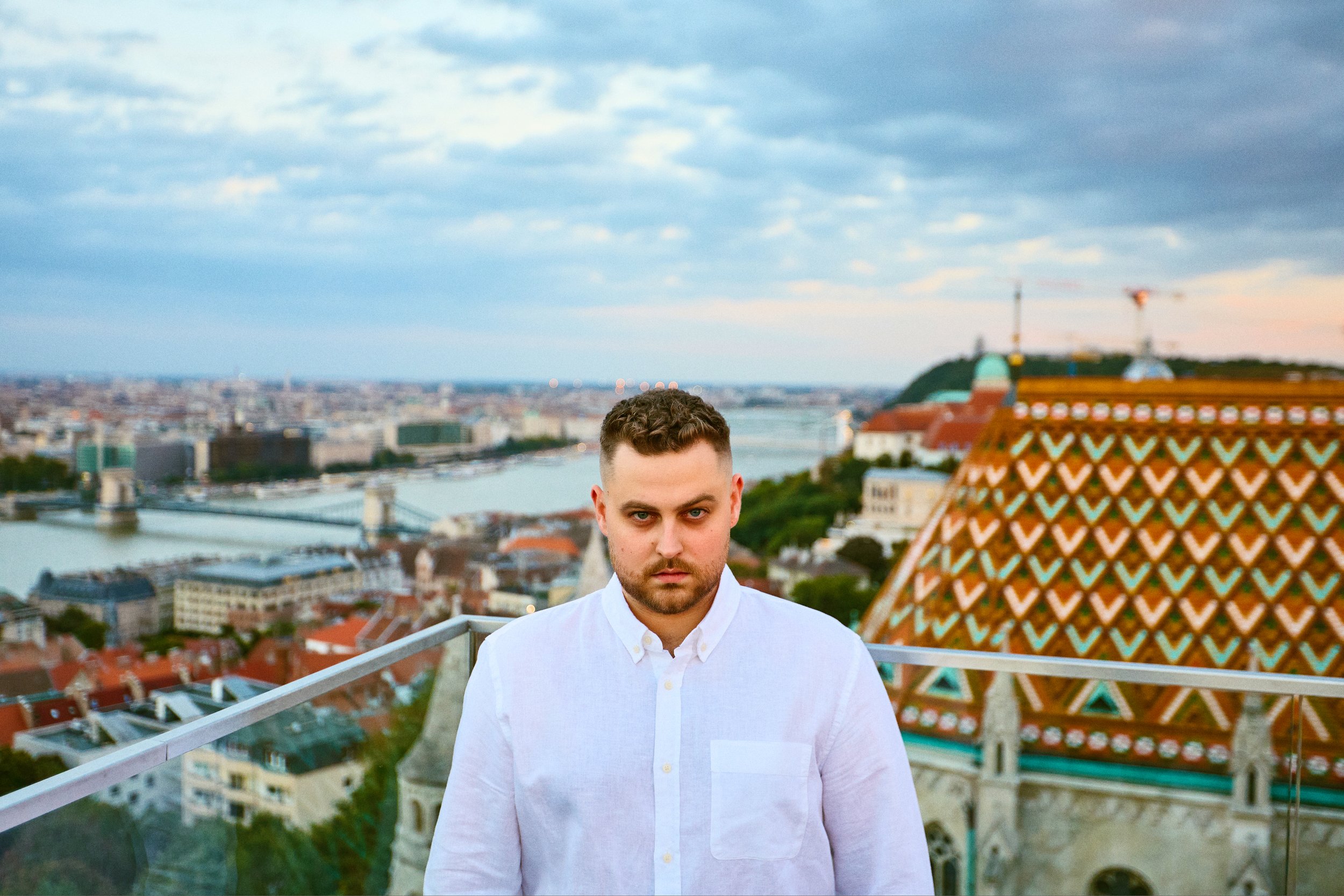 A man with short dark hair and a beard standing on a rooftop overlooking a cityscape with a river, historic buildings, and a bridge, during twilight.