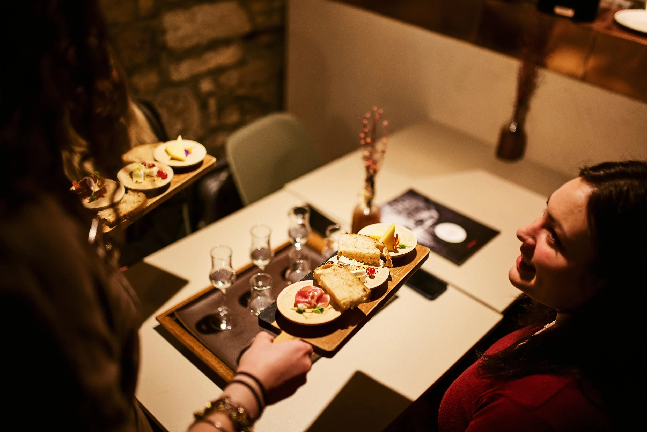 A woman serving a wooden tray with cheese, bread, and meats to another woman who is smiling, in a cozy restaurant setting.