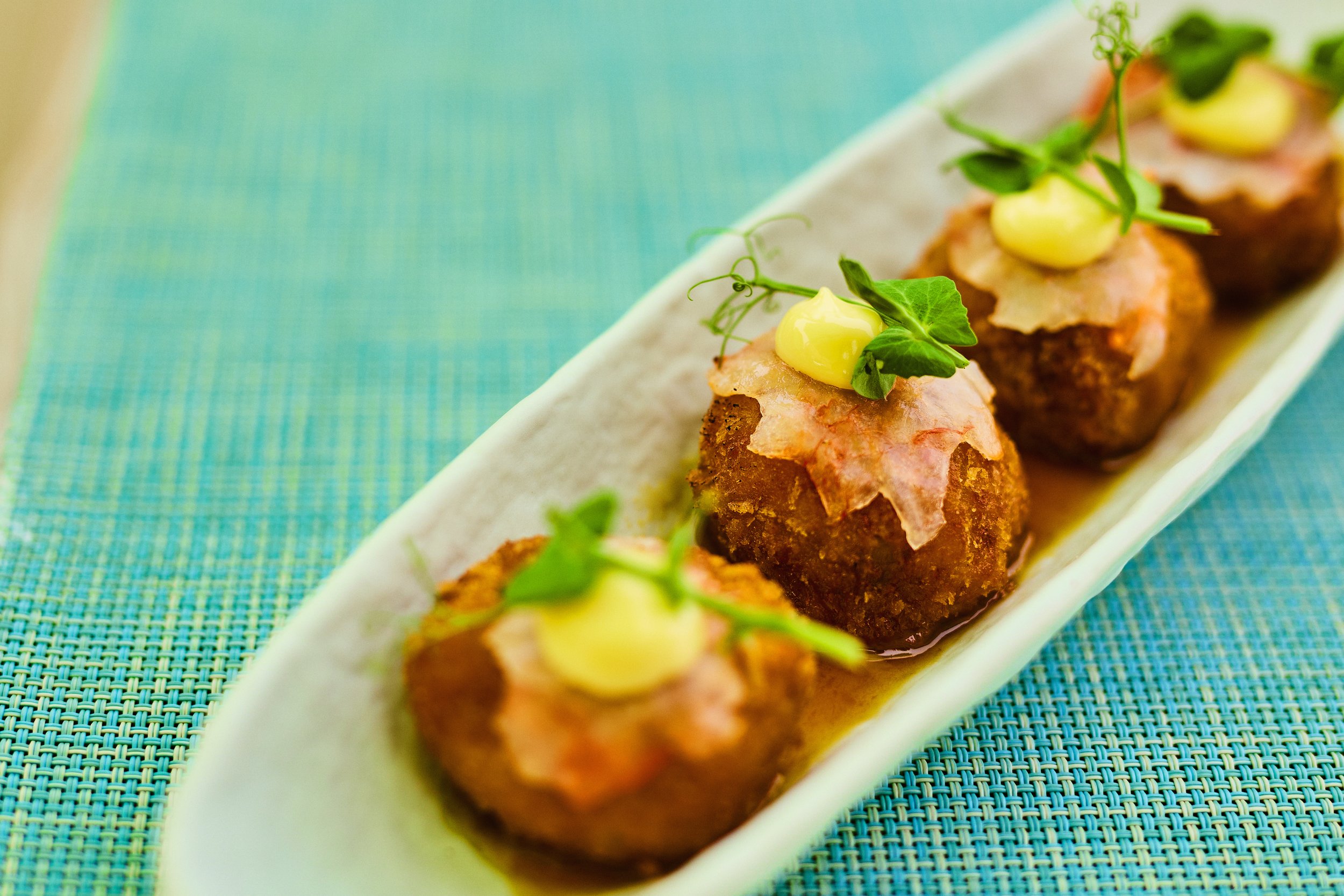 Close-up of four gourmet meatballs topped with a creamy yellow sauce, shredded ham, and green microgreens, served on a white rectangular plate with a colorful woven placemat.