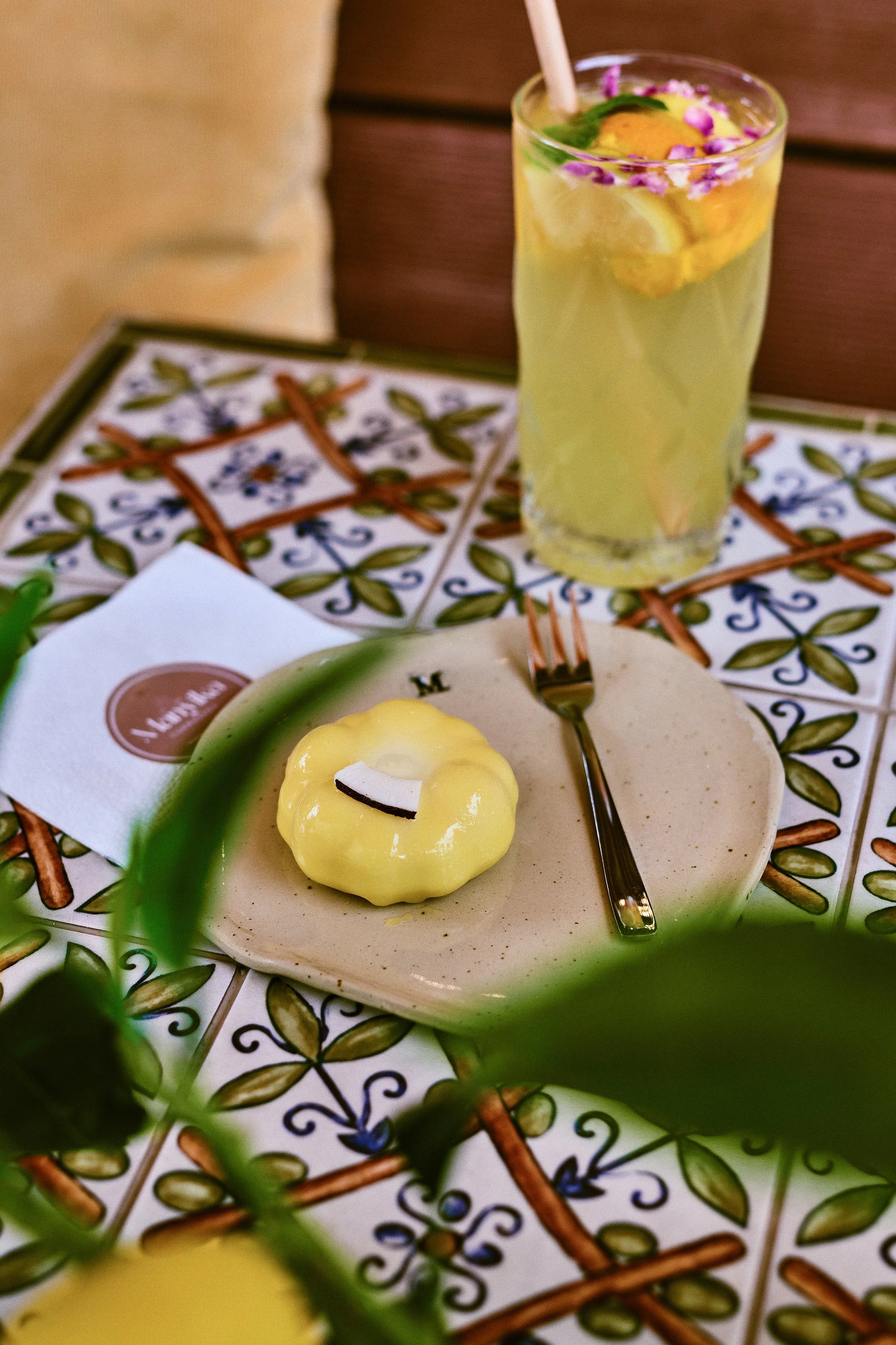 A yellow dessert with a small chocolate garnish on a beige plate, a fork beside it, and a tall glass of lemonade with lemon slices and purple flower petals on top, all on a decorative tiled table.
