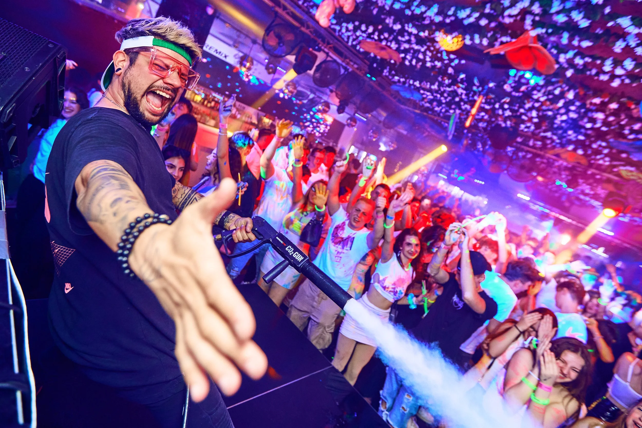 A DJ with tattoos, wearing pink sunglasses and a green headband, performing at a lively nightclub with a crowd dancing and colorful lights overhead.