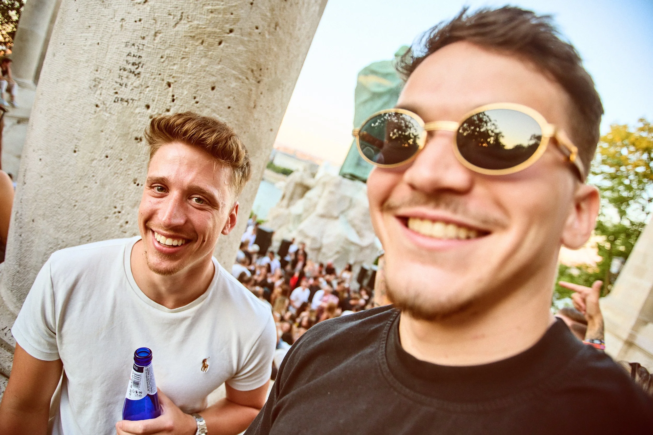 Two young men smiling at a social gathering outdoors. One is wearing a white t-shirt and holding a beer, the other is wearing sunglasses and a black shirt. There are many people in the background.