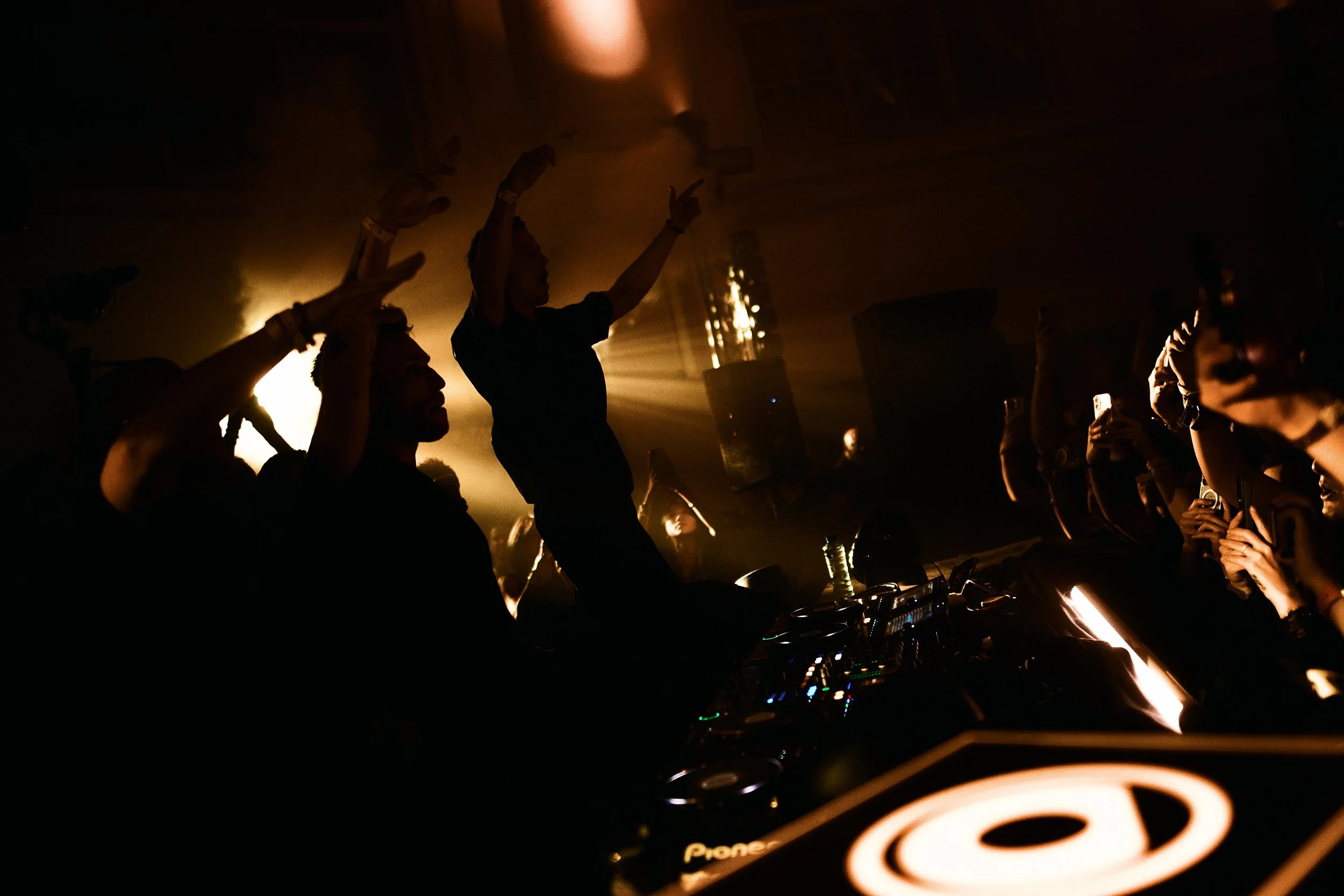 Silhouettes of people dancing and enjoying music at a nightclub or concert with DJ booth and colorful lighting.
