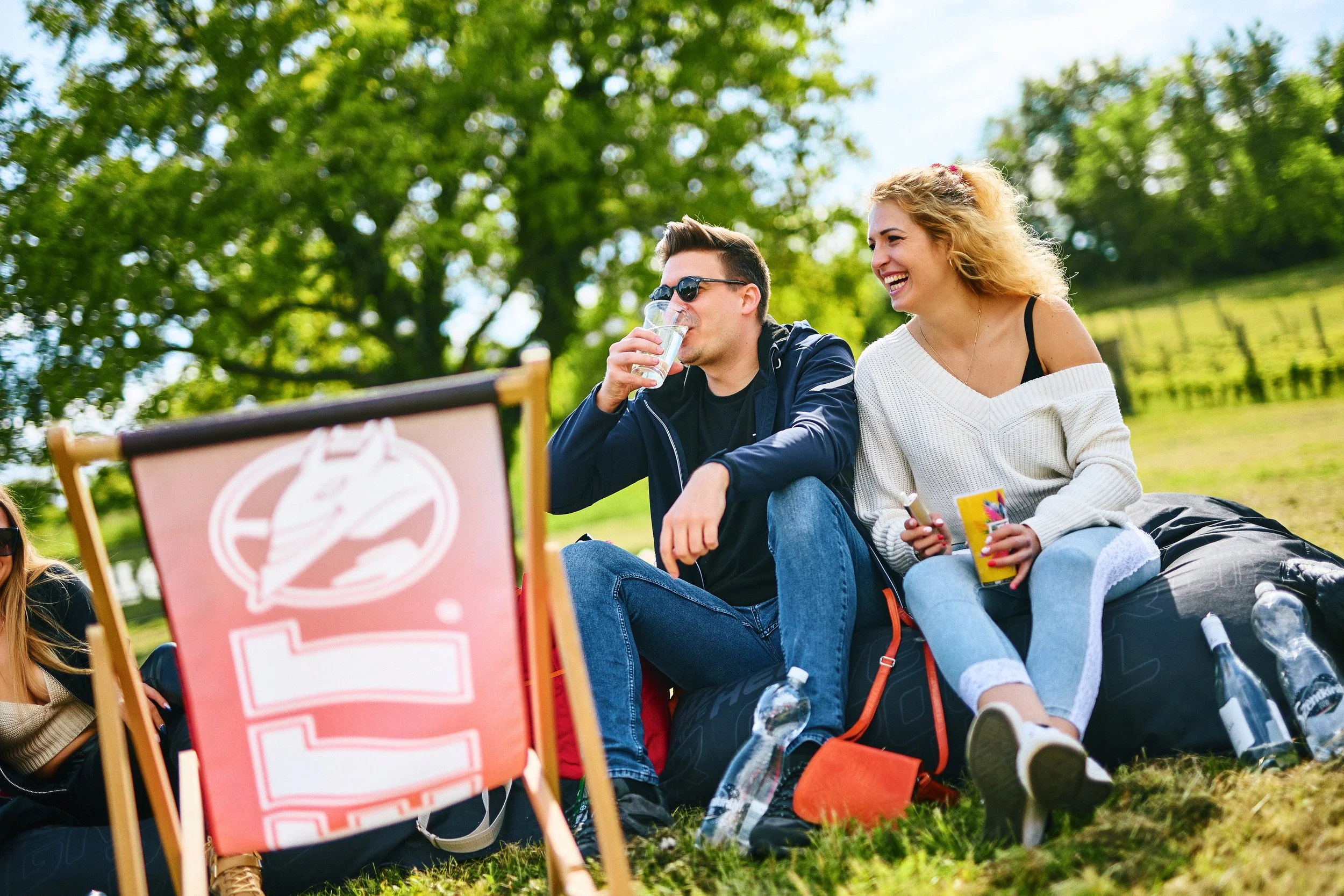 Two young adults, a man and a woman, sitting in a grassy outdoor area on a sunny day, enjoying snacks and drinks, with trees and a fence in the background.