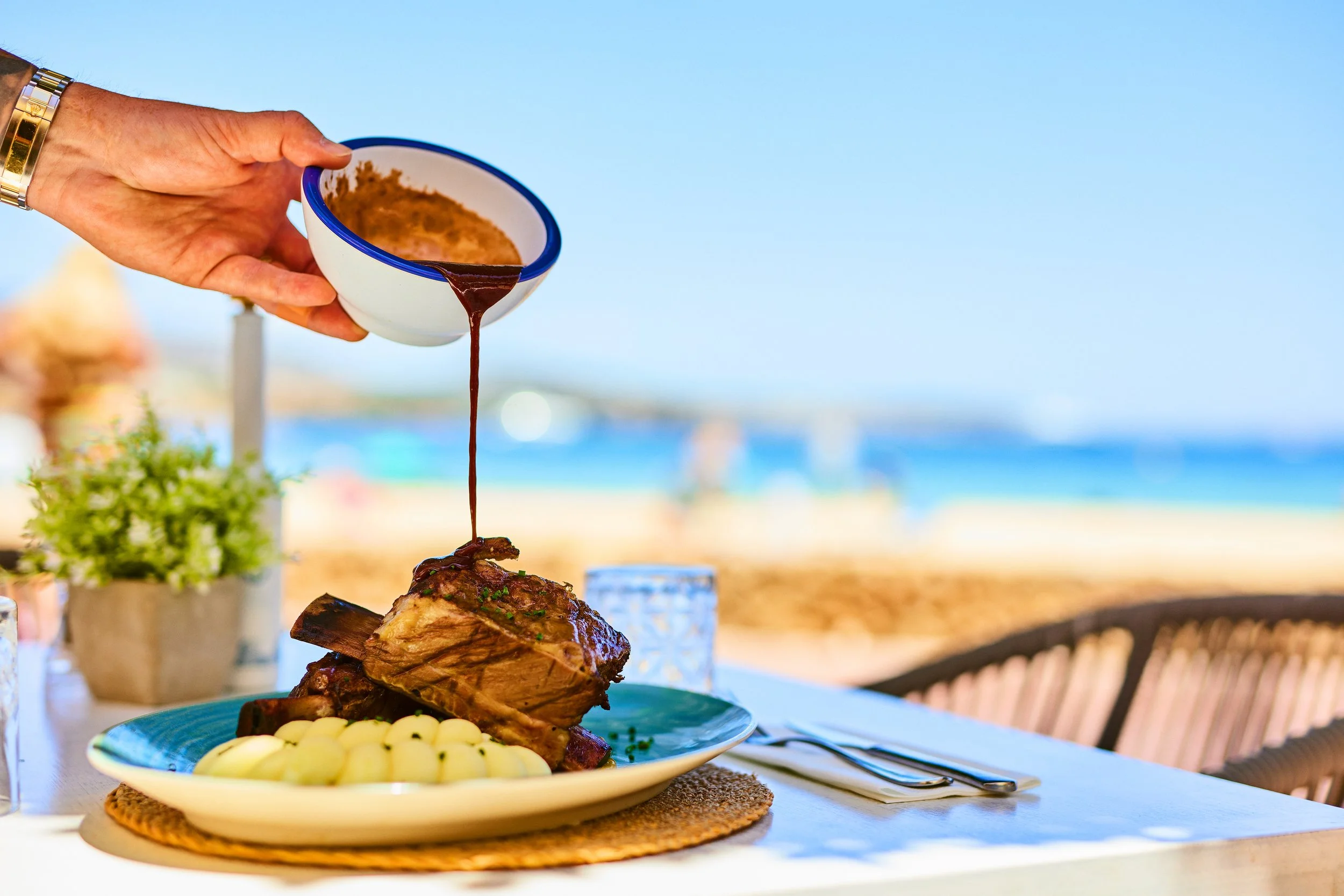 A person drizzling gravy over a large piece of cooked meat with potatoes on the side, set on a table outdoors near a beach.