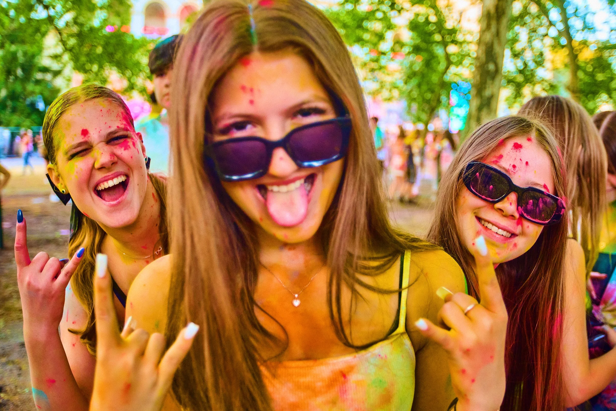 Three young women at a colorful outdoor celebration covered in vibrant powder, wearing sunglasses, smiling and making rock and roll hand gestures.