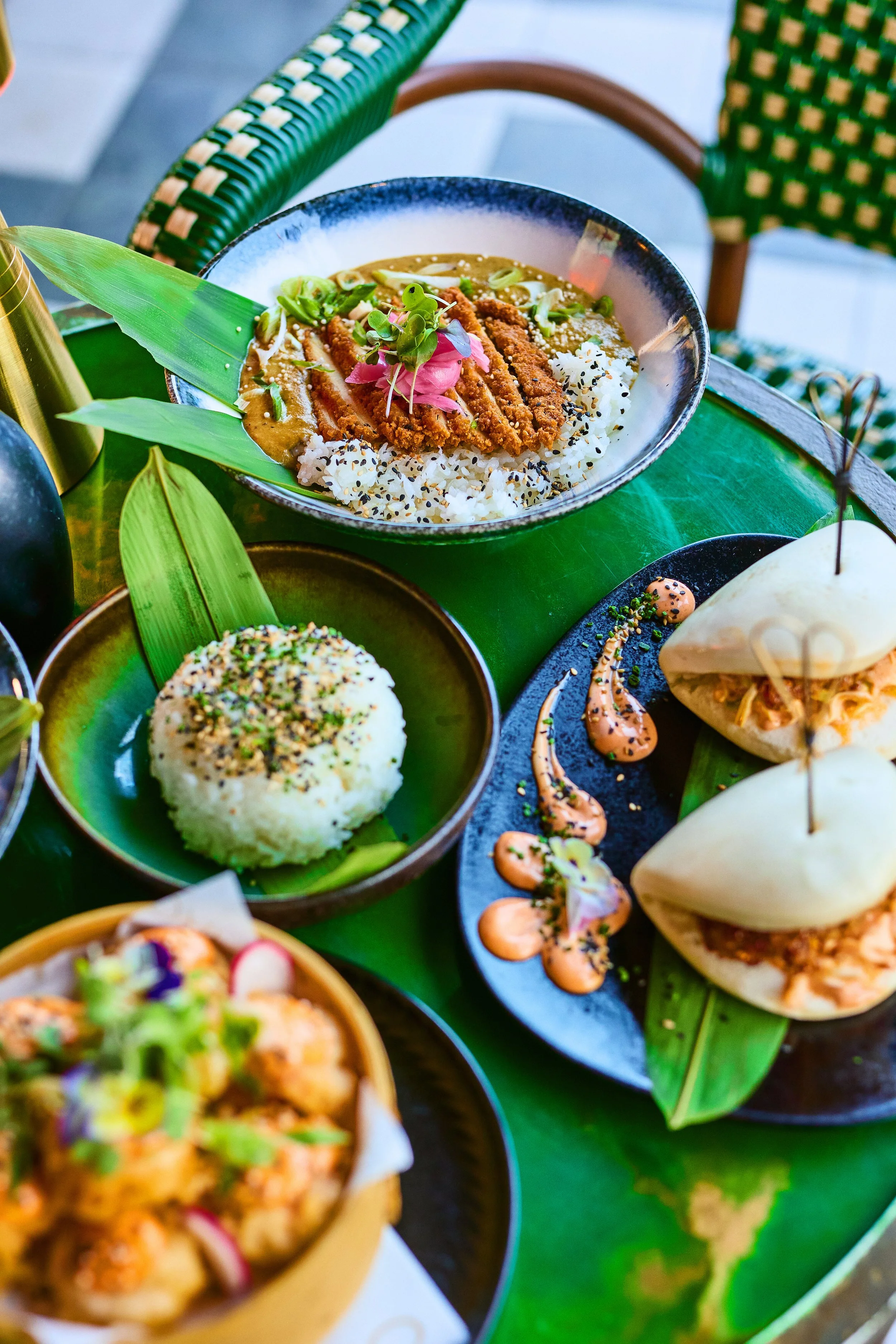 Assorted dishes including rice, curry, and steamed buns served on green leaves and black plates on a green table.
