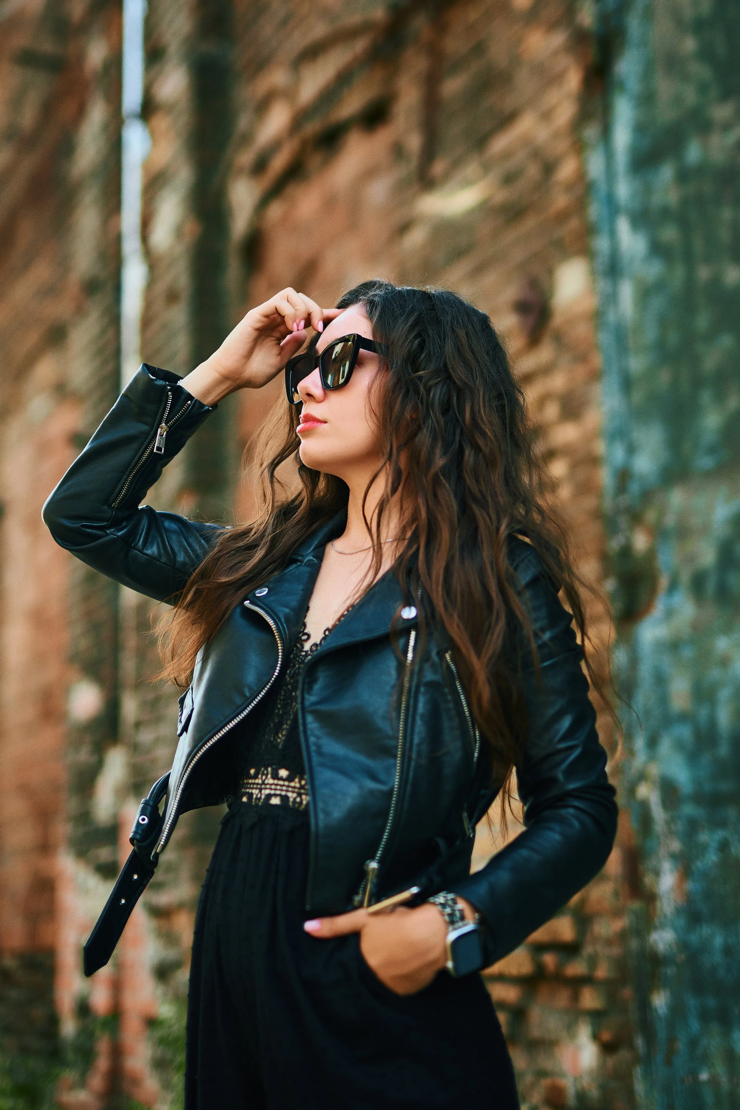 A woman with long wavy brown hair wearing black sunglasses, a black leather jacket, and a black dress standing outdoors against a brick wall.