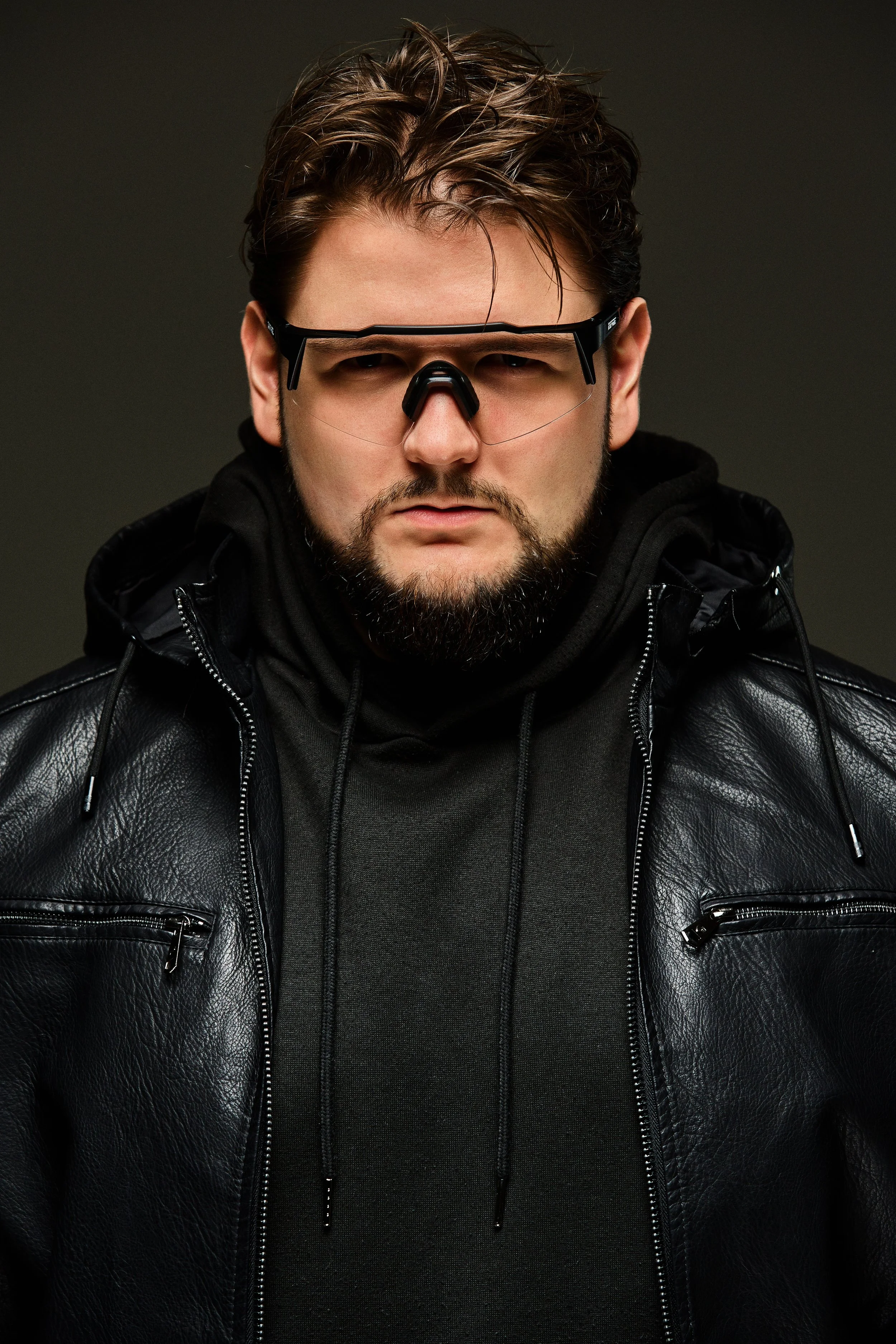 A man with dark hair and a beard wearing black sunglasses, a black hooded sweatshirt, and a black leather jacket.