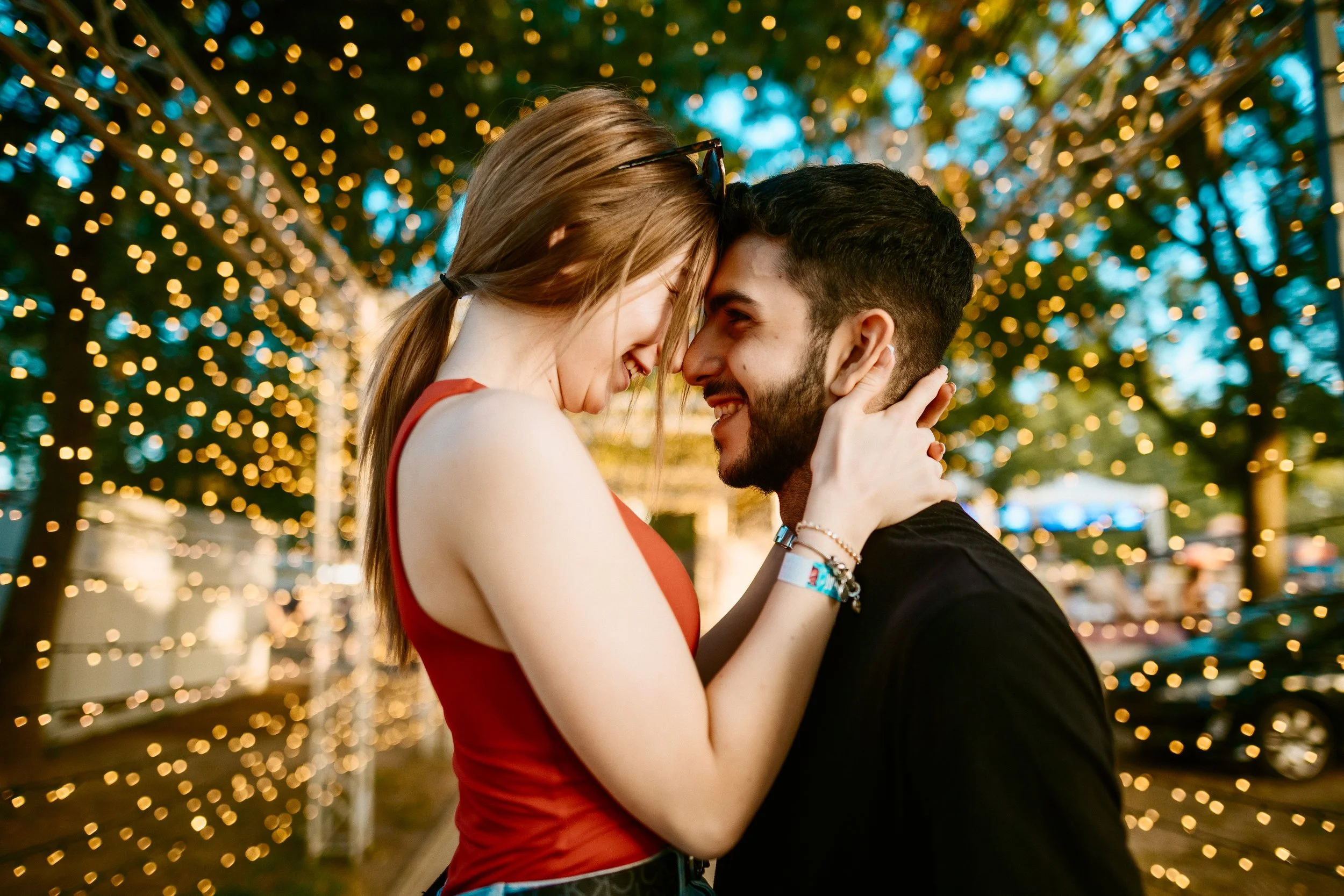 A young couple is close together, smiling and touching foreheads, outdoors at night with string lights in the background.