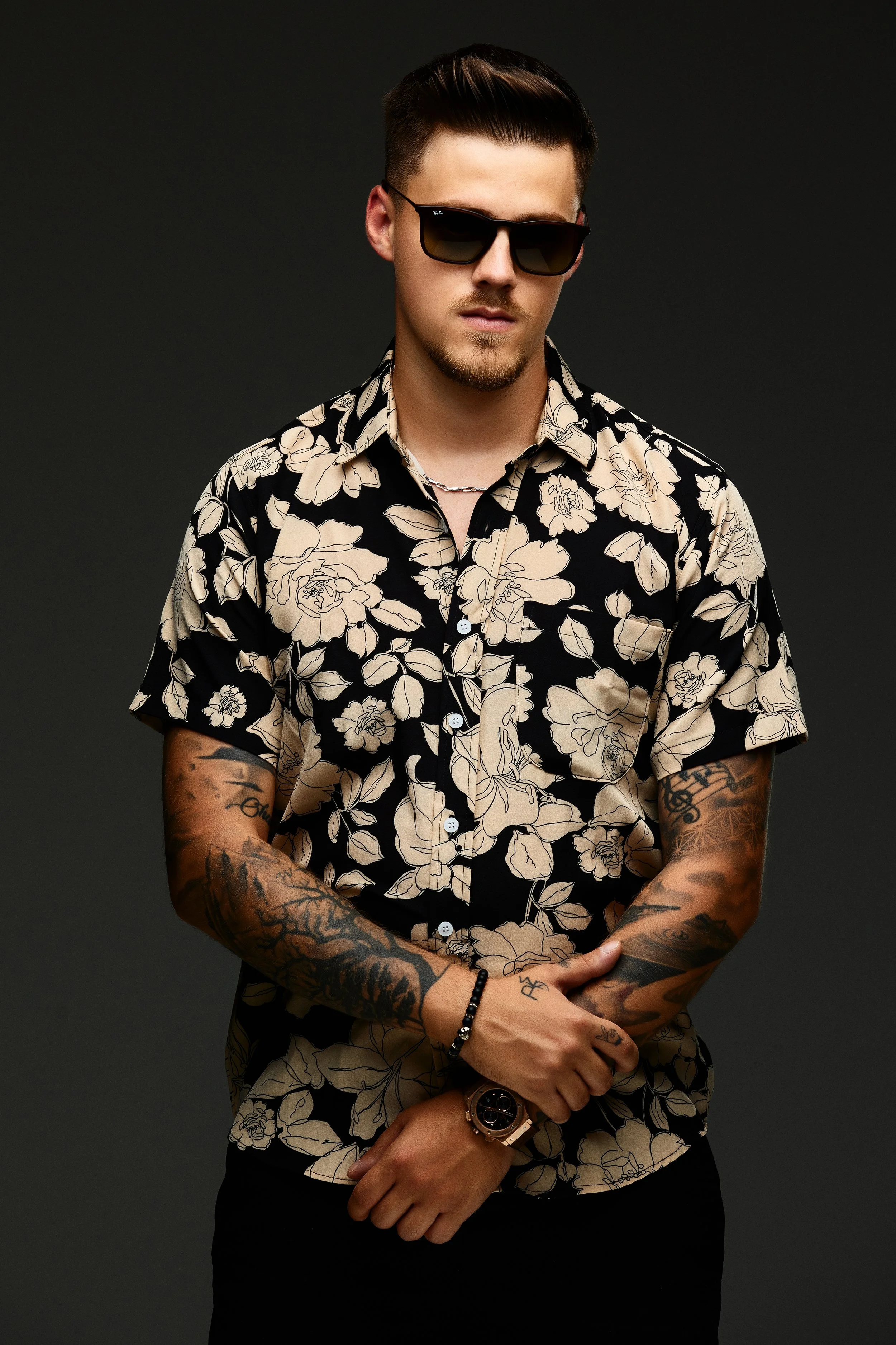 A young man with tattoos, wearing sunglasses and a black and beige floral shirt, standing against a dark background.