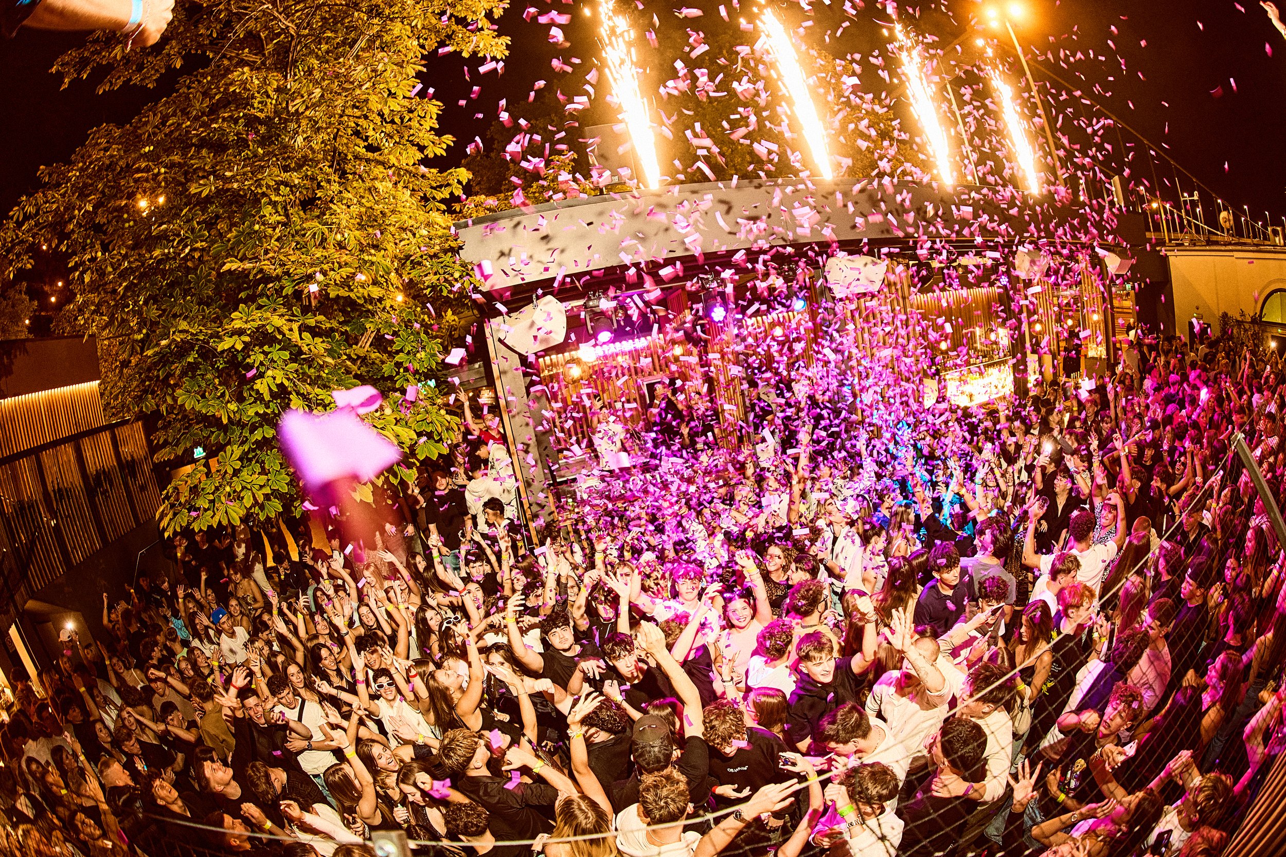 A lively outdoor party with a large crowd of people celebrating as colored confetti falls from the sky and fireworks explode overhead. The scene is set at night with festive lighting and a stage in the background.