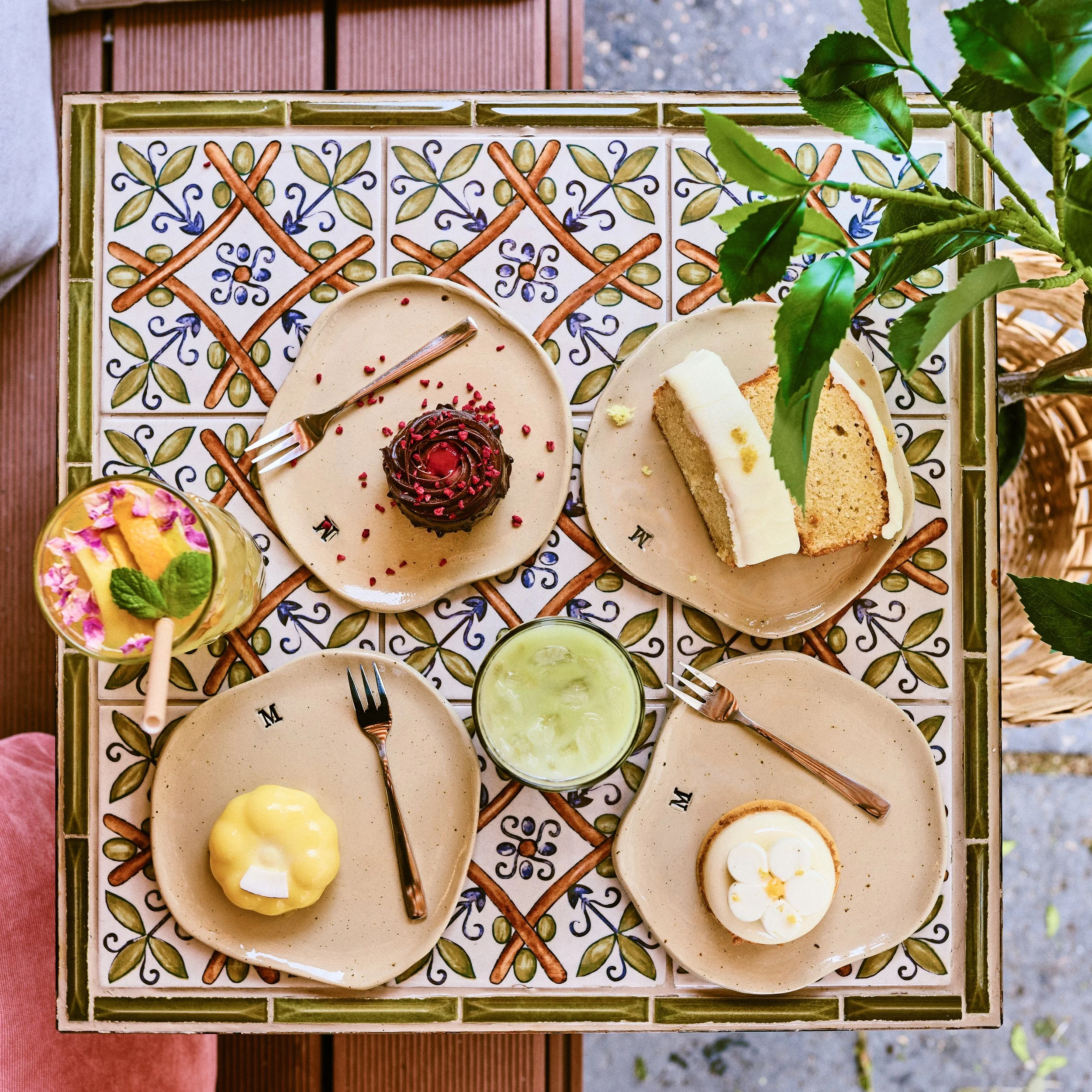 Top-down view of a colorful dessert and cake platter on a decorative tiled table with plant leaves in the corner.