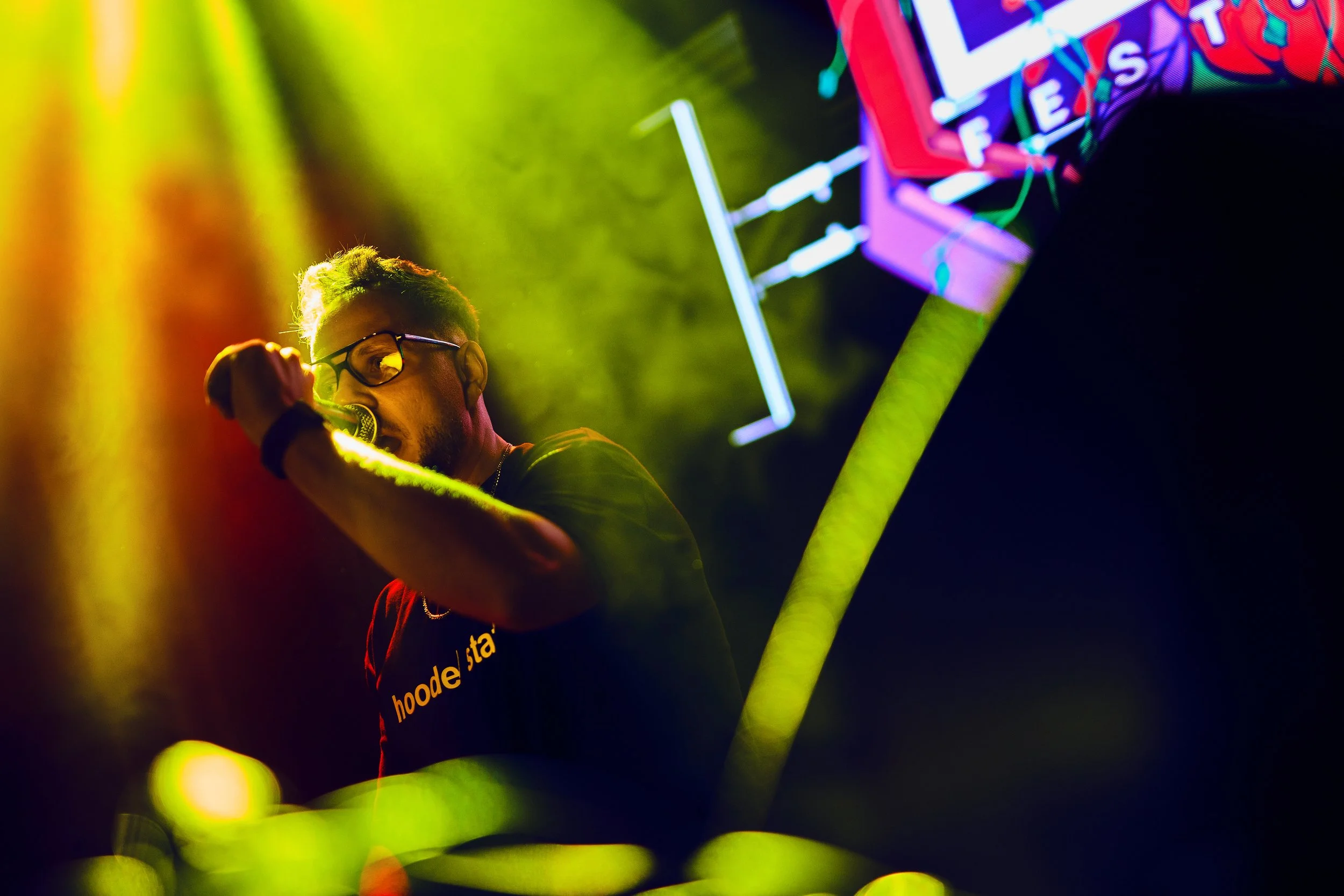 A person wearing glasses and a black t-shirt with yellow text, performing at a neon-lit event, surrounded by colorful lighting and a decorative neon sign.