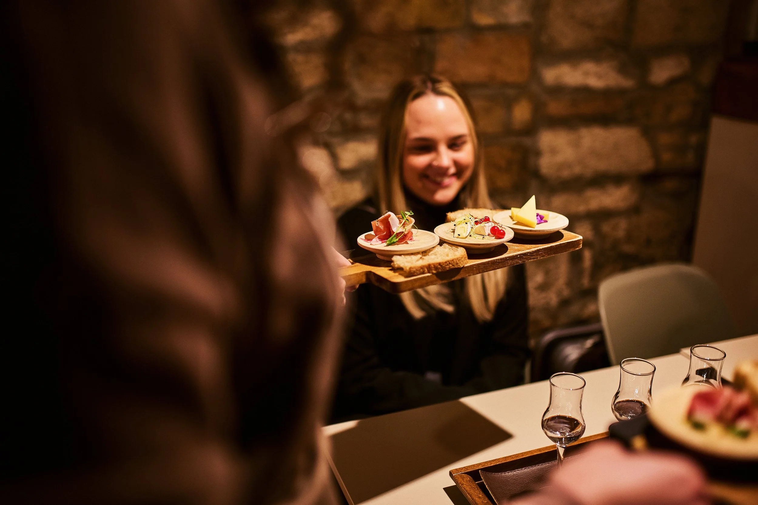A woman smiling while holding a tray of assorted small dishes with cheese, meats, and garnishes in a restaurant with a brick wall background.