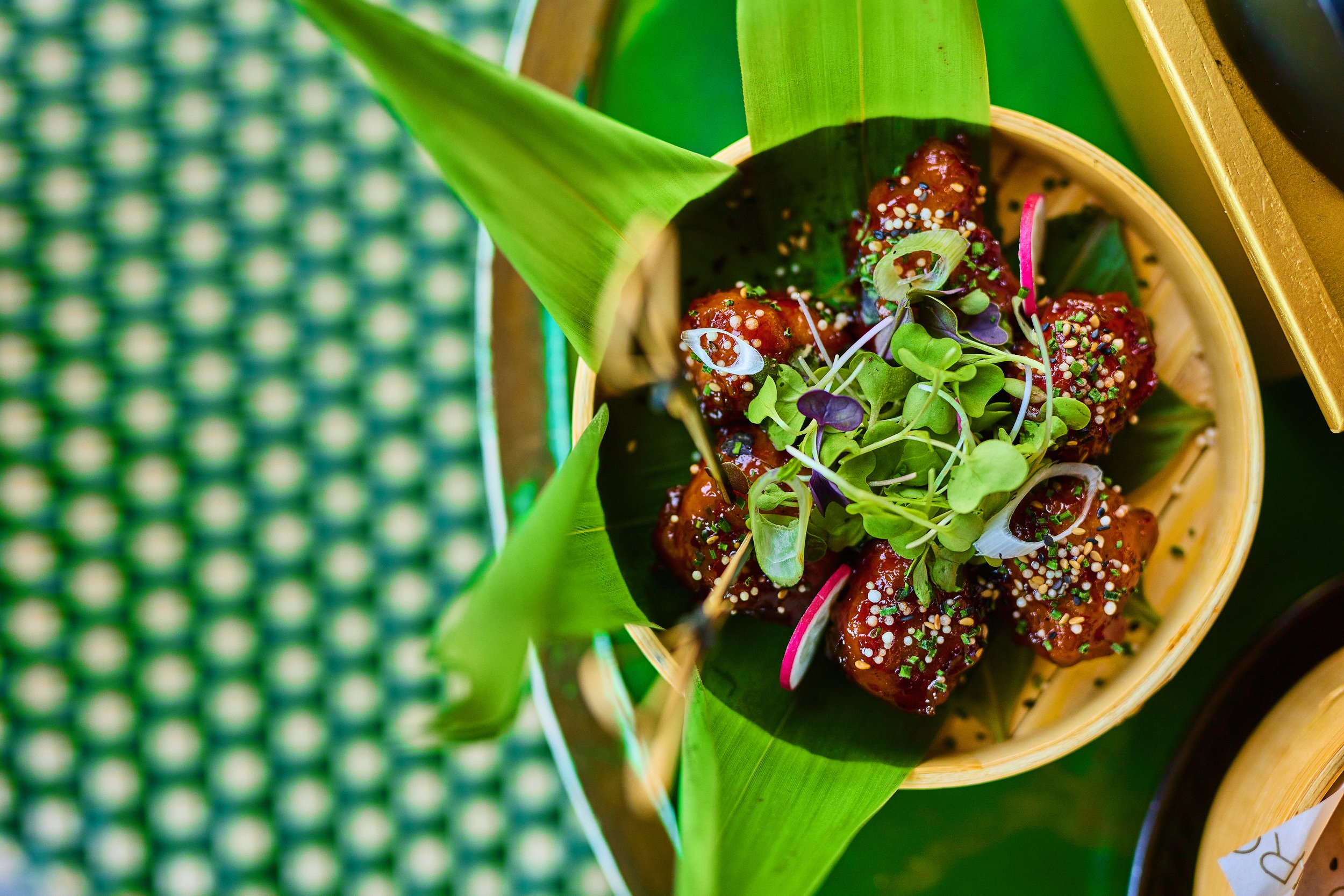A bamboo steamer basket with spicy glazed chicken pieces topped with green microgreens and sesame seeds, on a green leaf mat.