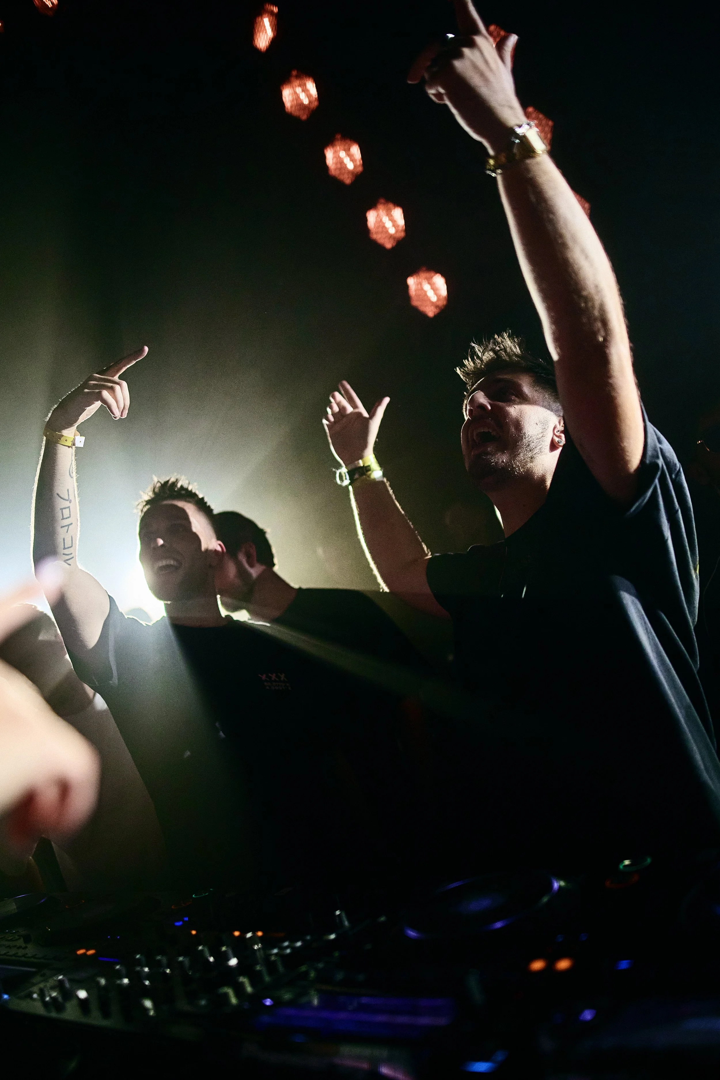 Two men dancing at a nightclub with dim lighting, colorful lights, and a DJ mixer in the foreground.