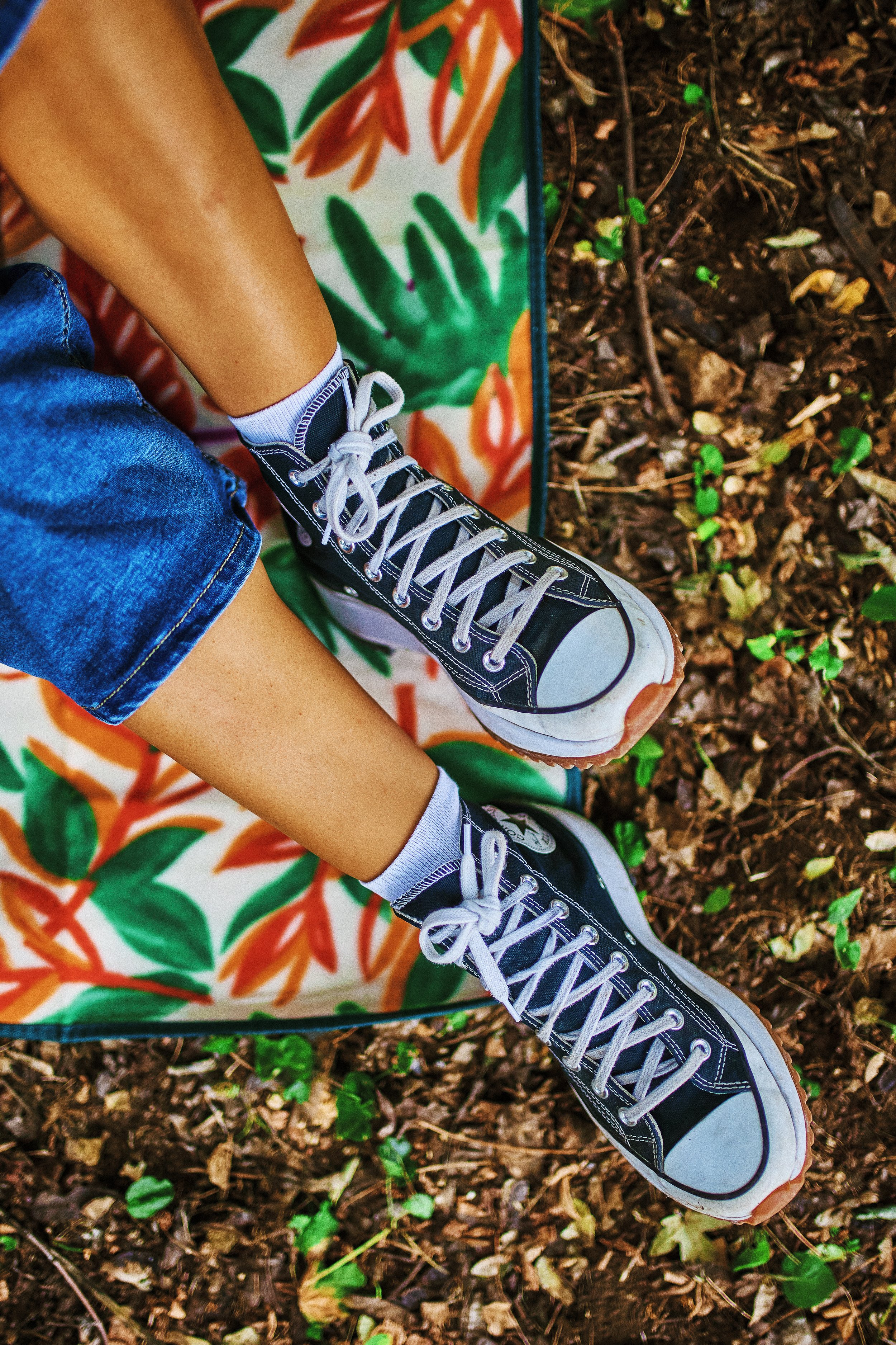 Person wearing black high-top Converse sneakers, blue jeans, and white socks with black accents, sitting on a colorful blanket with a floral pattern on a bed of leaves and small plants.