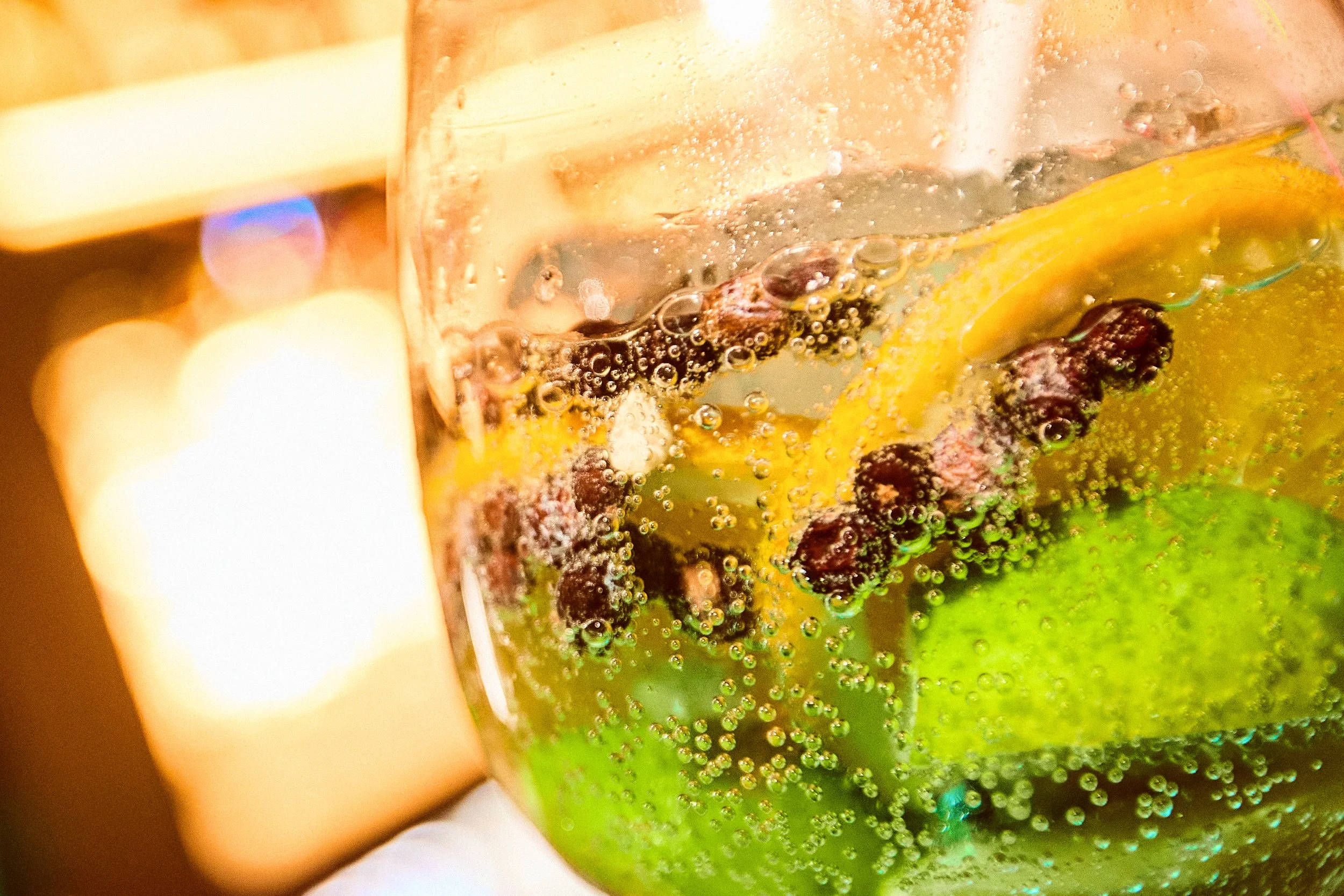 Close-up of a clear glass filled with bubbly soda containing slices of lemon and lime, with black peppercorns floating inside.