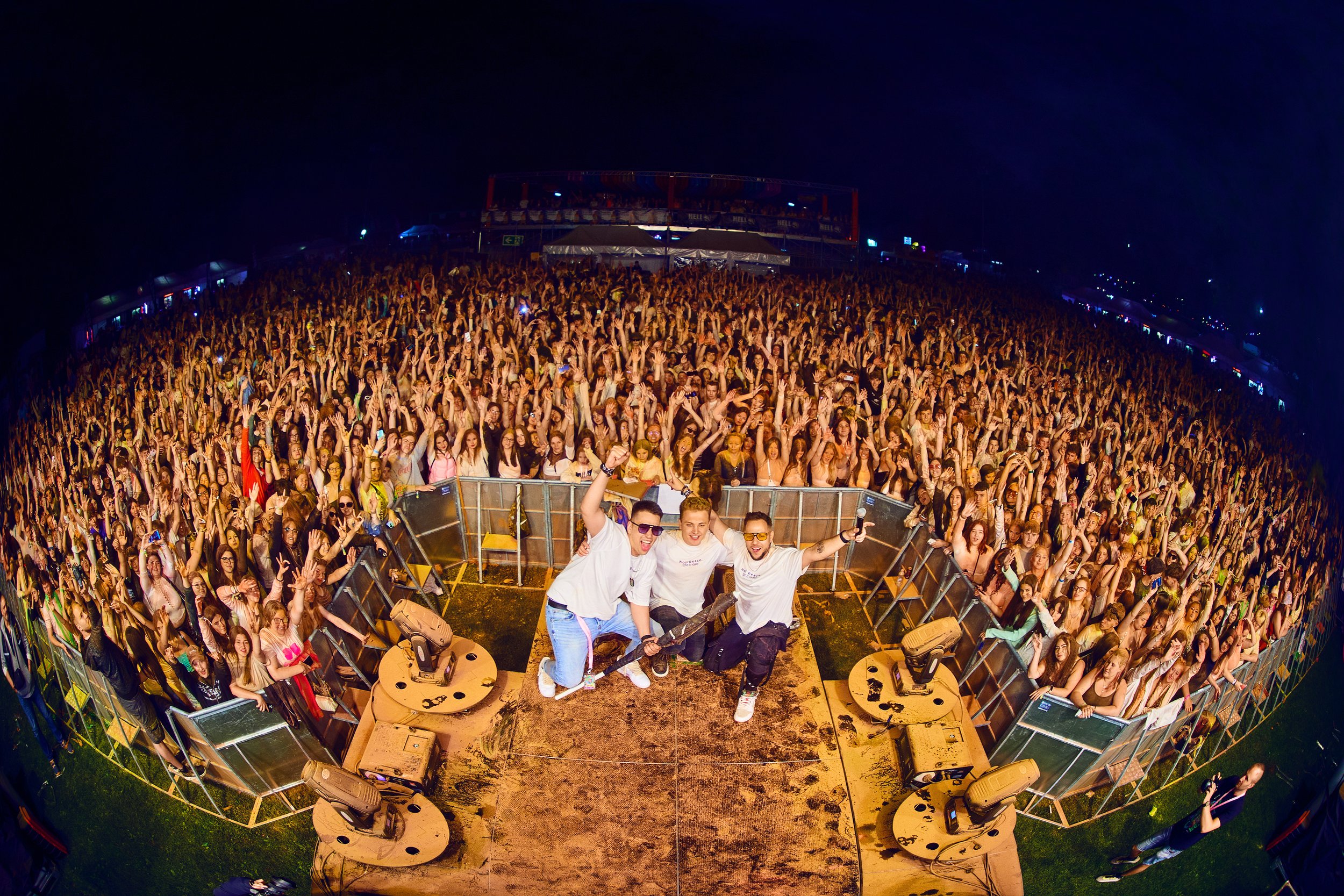 Four performers on stage at an outdoor concert, with a large crowd of cheering fans behind them at night.