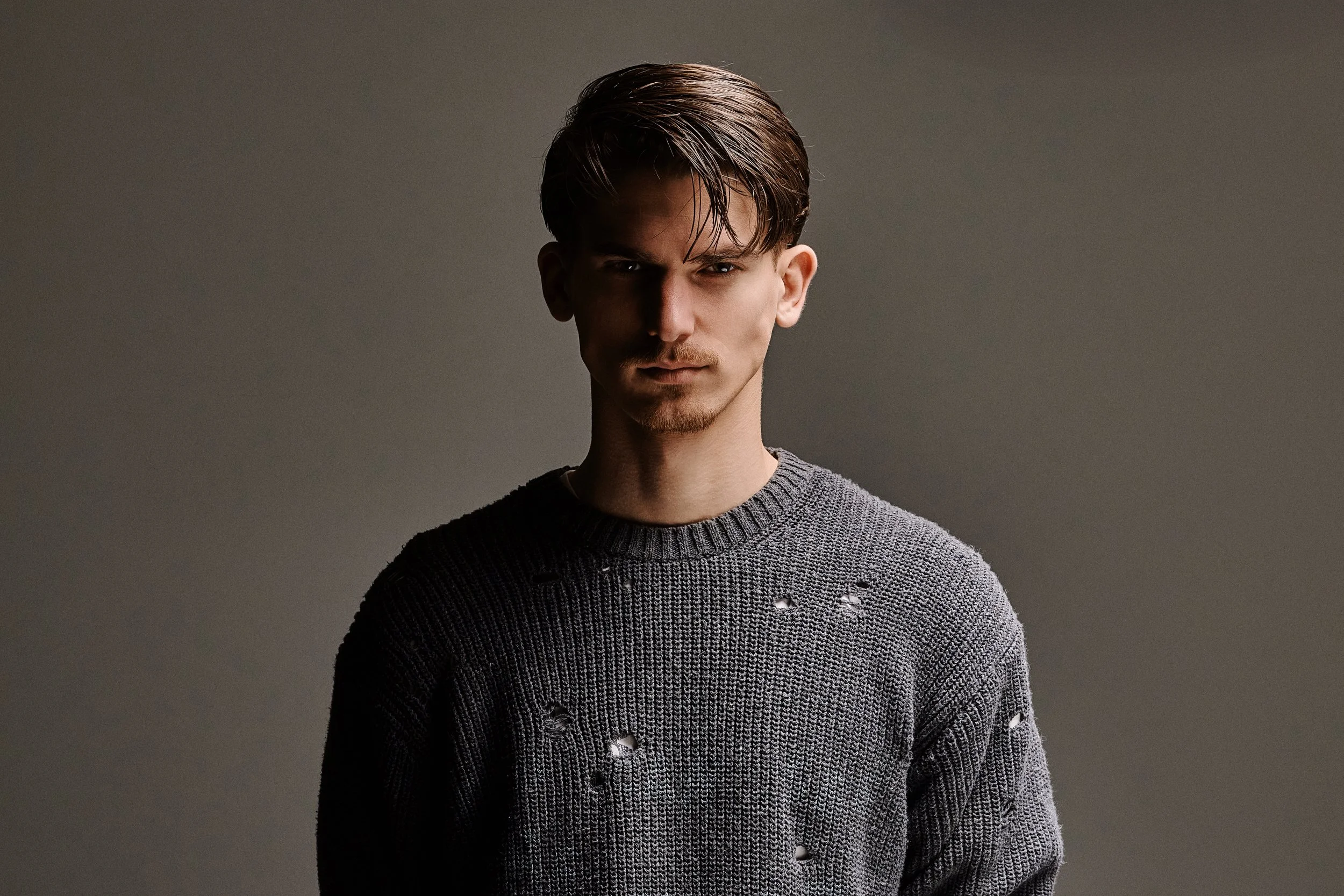 A young man with dark brown hair styled back, wearing a distressed gray sweater, standing against a plain gray background.