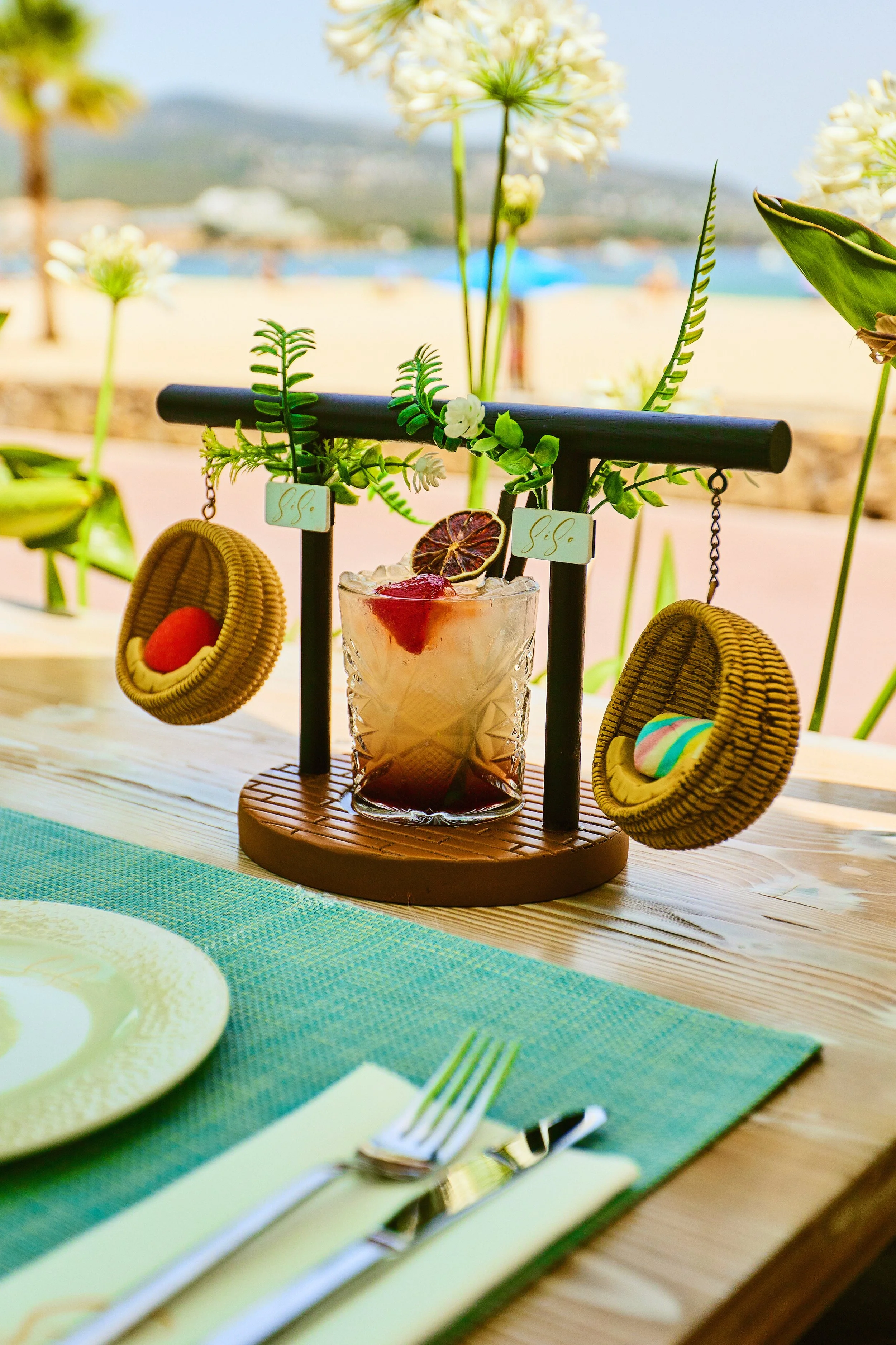 Colorful cocktail with fruit garnish on a decorative wooden drink holder, placed on a wooden table with a green placemat, outdoor beach background with flowers and palm trees.