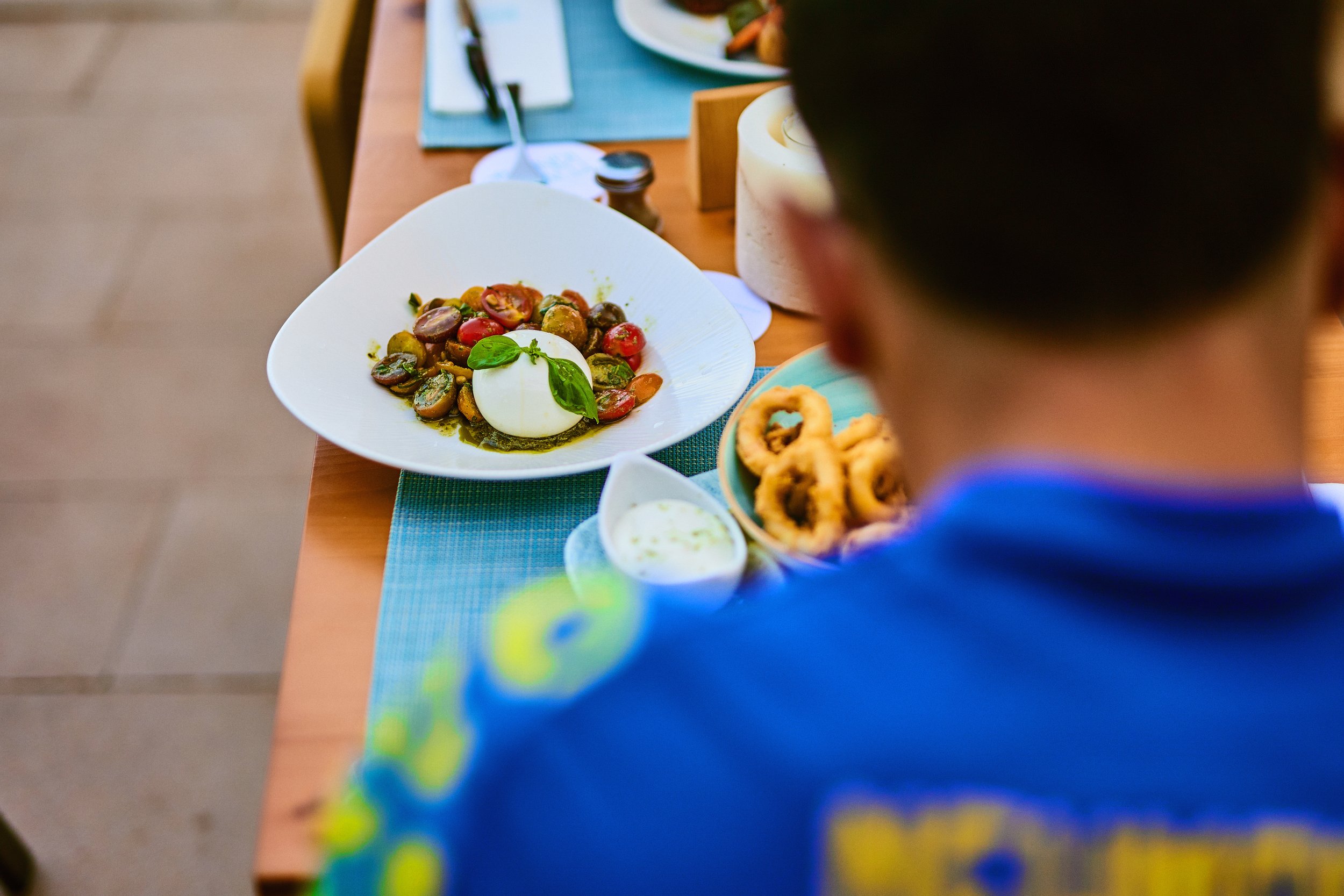 Person in blue shirt sitting at a table with a plate of mixed cherry tomatoes and a ball of mozzarella cheese topped with basil, alongside a bowl of fried calamari and a small dish of dipping sauce.