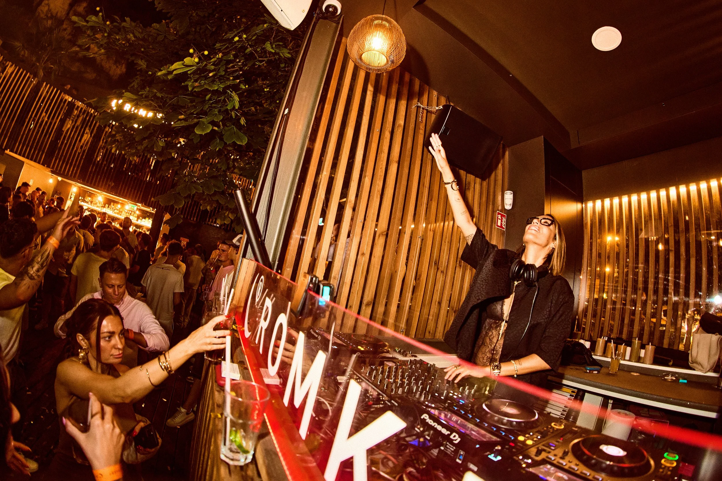 A female DJ with short hair wearing glasses and a lace top, raising her hand while performing at a crowded indoor party with a wooden wall and decorative lighting.
