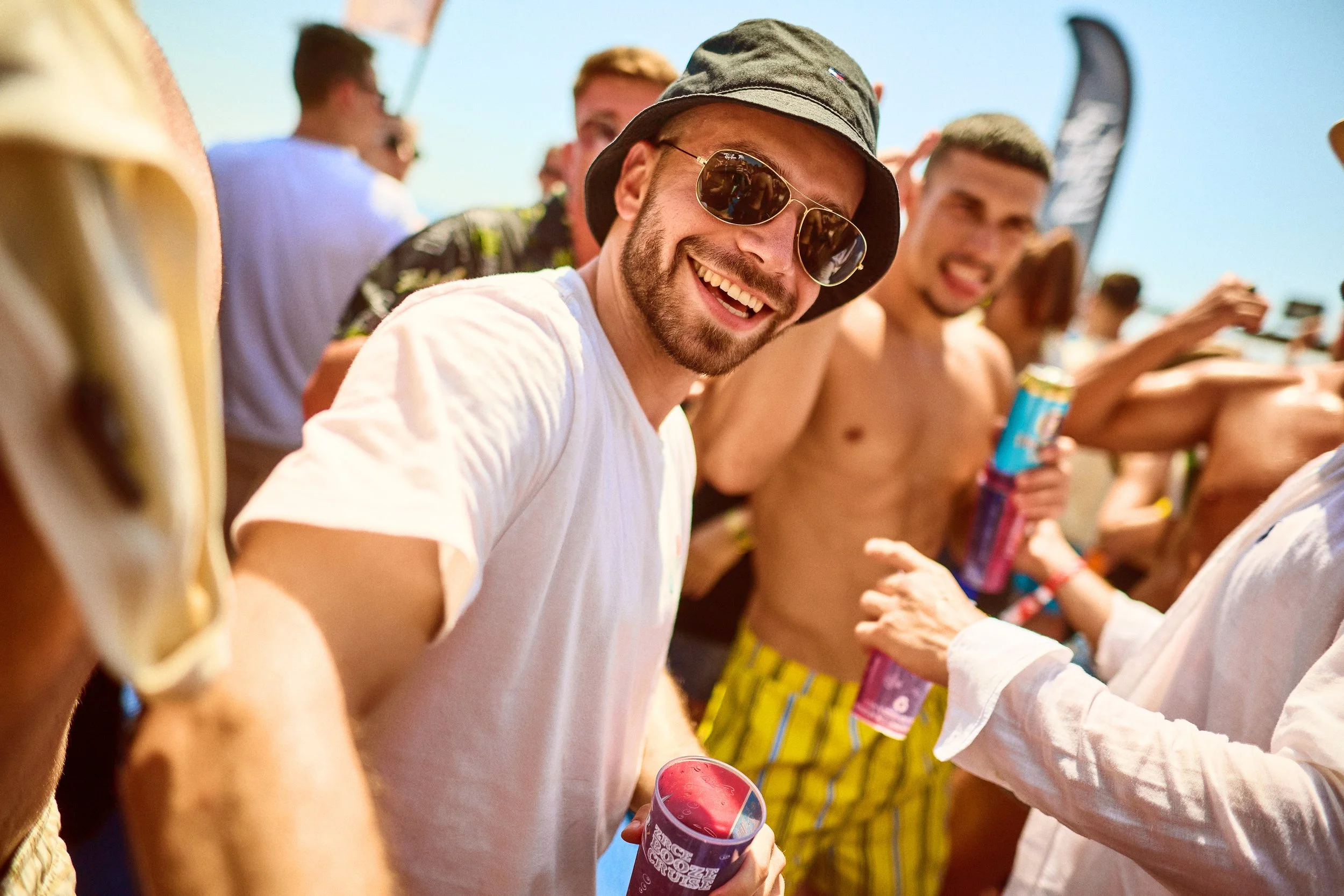 Groups of people at an outdoor event enjoying the sun, some shirtless, holding drinks, with tents and flags in the background.