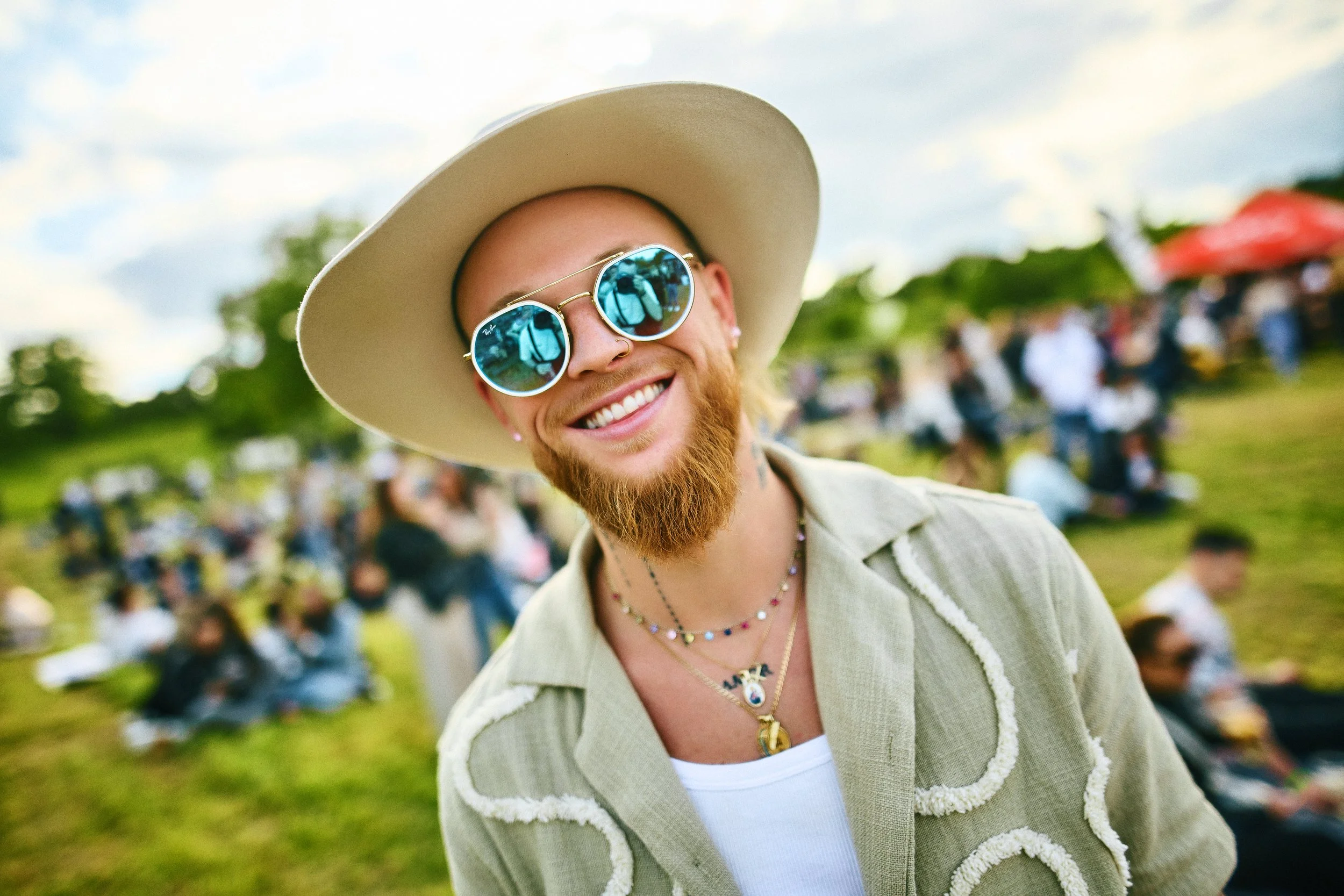 Smiling man with a red beard wearing sunglasses, a wide-brimmed hat, and layered necklaces at an outdoor event with a crowd and tents in the background