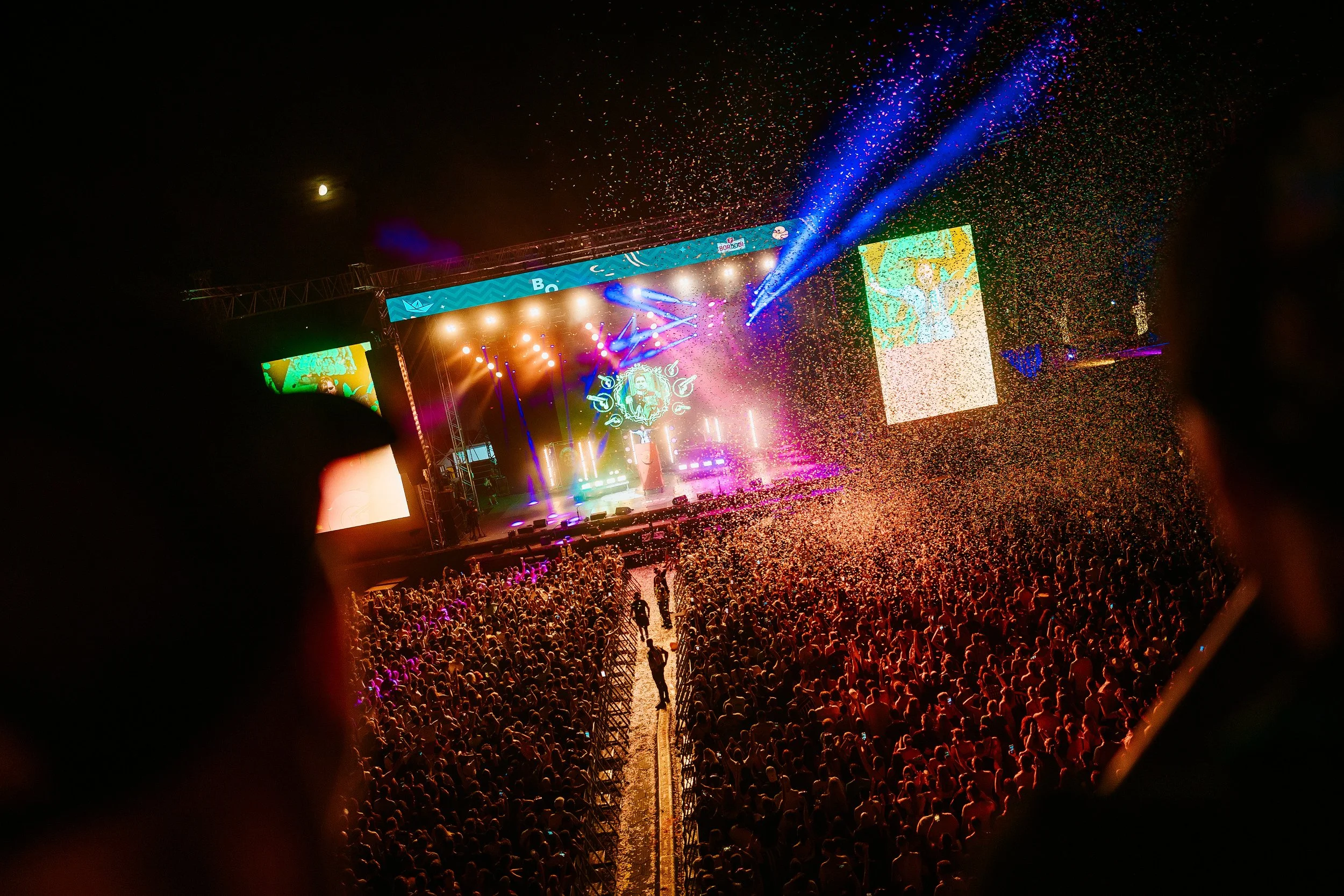Night concert scene with a large crowd, colorful stage lights, and confetti falling during a music performance.