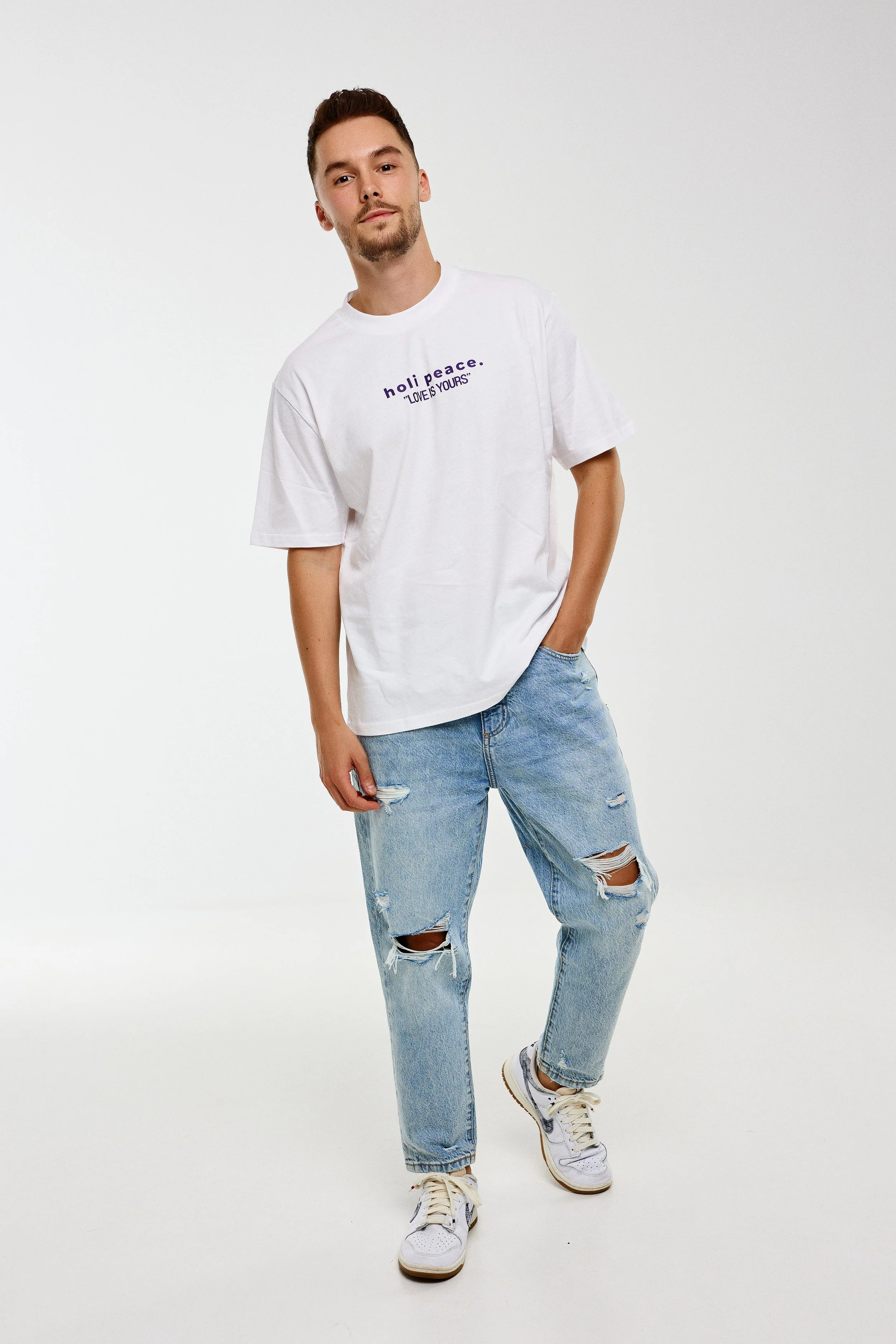 A young man wearing a white t-shirt with the text 'holl peace. LOVE YOURS' and distressed blue jeans with rips at the knees, standing against a plain white background.
